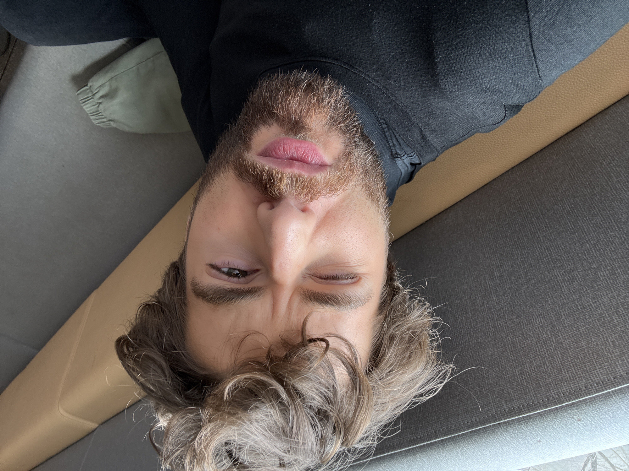 The image is an upside-down selfie of a man with light brown hair and a beard. He is lying down, winking with his right eye, on a gray couch with a tan armrest. He is wearing a black top. The location is unknown, and there is no verifiable indication of Montreal, Canada.