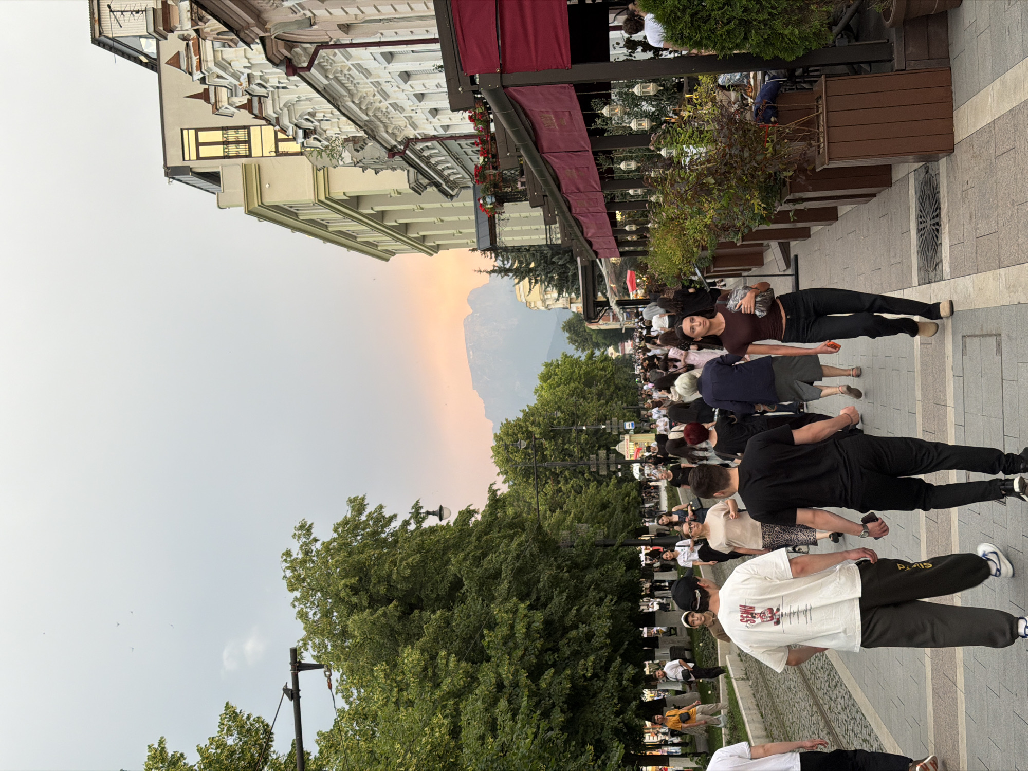 This image depicts a bustling street scene in Verkhniy Alkun, Russia. The street is lined with trees on the left and buildings with awnings and outdoor seating on the right. Numerous people are walking in both directions on the pedestrian walkway. The sky exhibits a soft sunset glow with visible mountains in the distance. People are dressed in casual summer attire. The general atmosphere suggests a lively urban environment, possibly a commercial or tourist area.