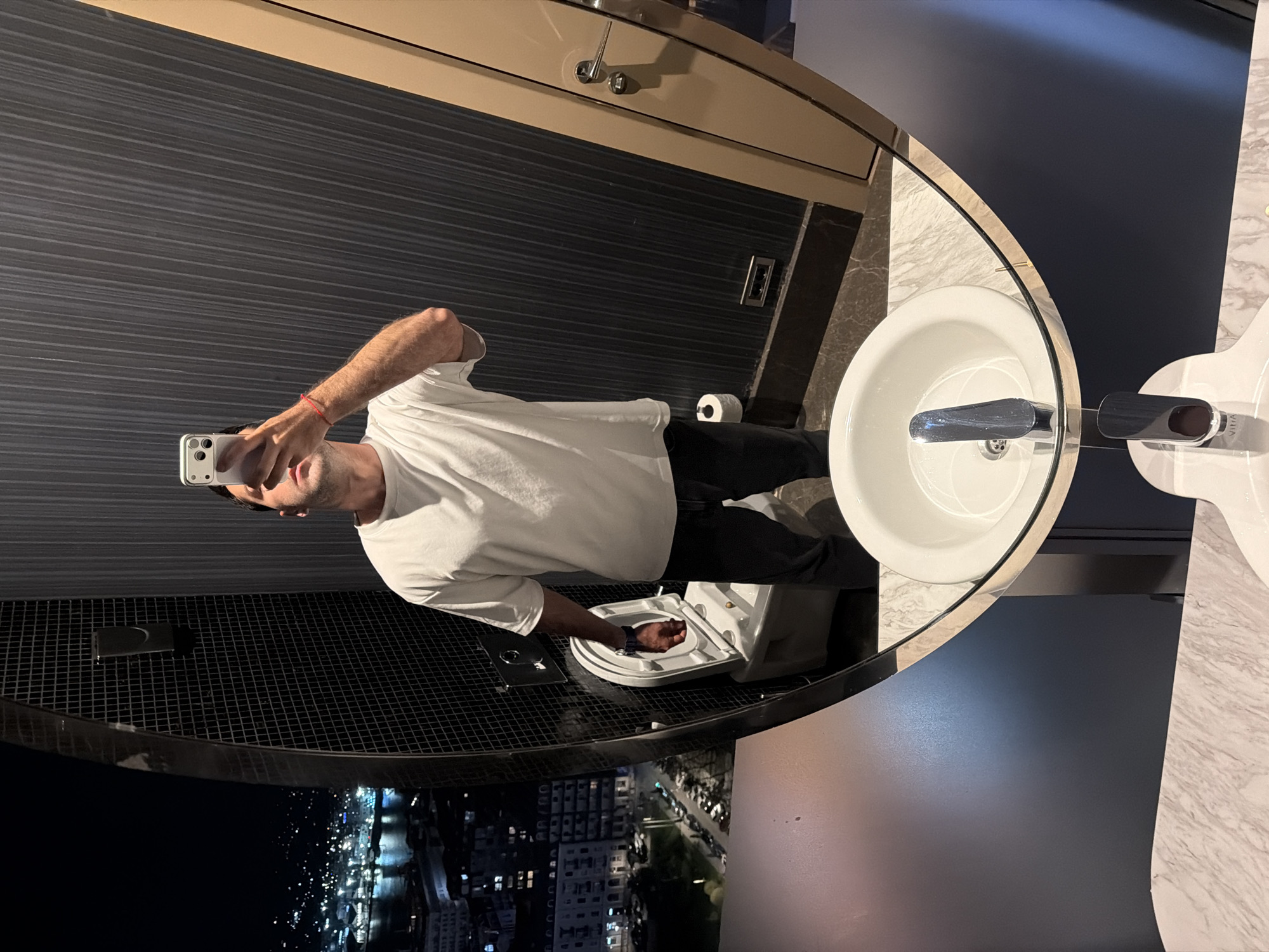 A selfie is being taken in a bathroom of a high-rise building, likely a hotel, in Batumi, Georgia, at night. Reflected in an oval mirror is a man wearing a white t-shirt and black pants. He is holding a white smartphone with his right hand. Behind the man is a toilet; to the right is a white countertop sink. Out the window is a view of the city illuminated at night.