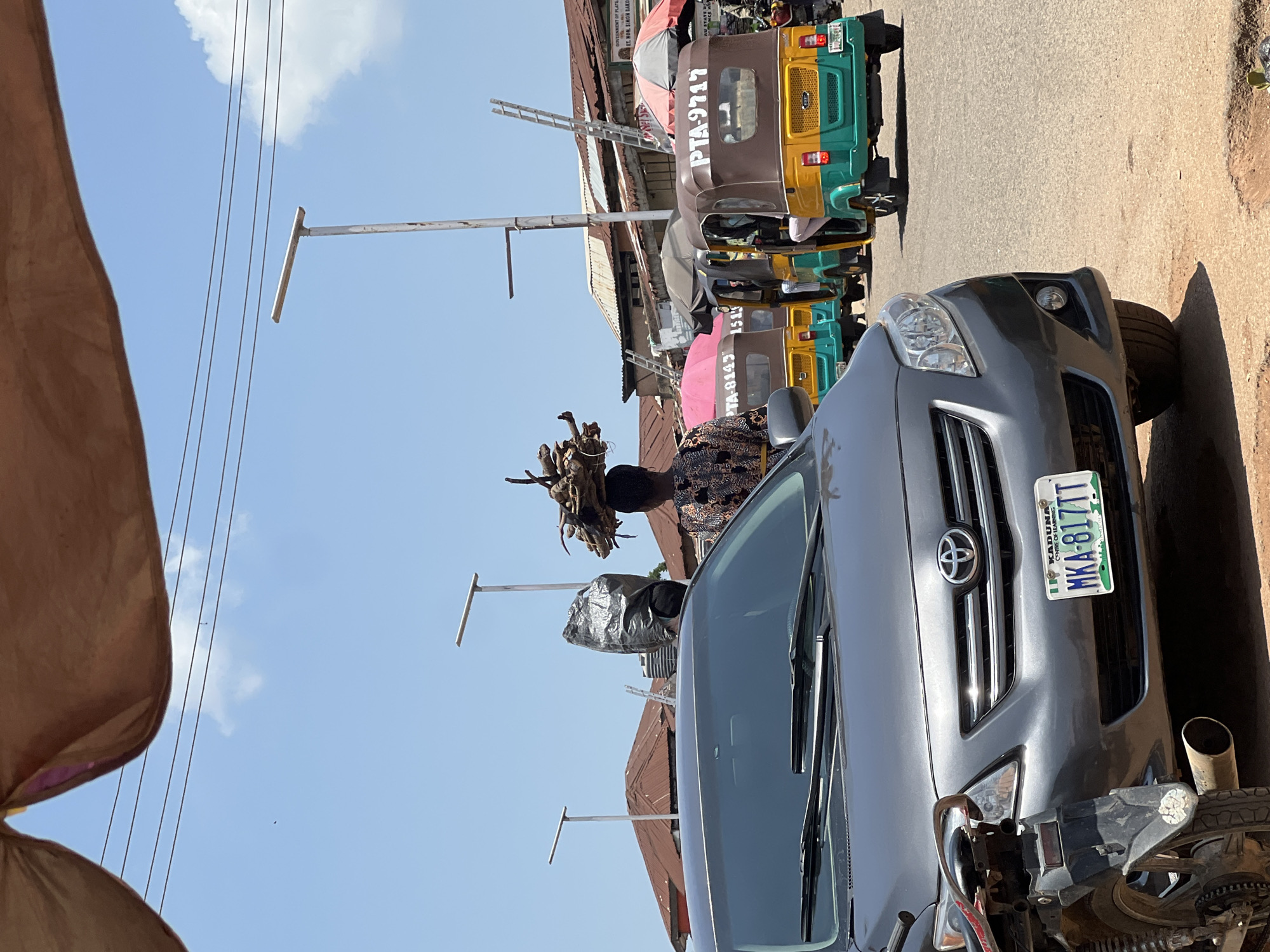 A medium shot captures a street scene in Jos, Nigeria, dominated by a gray Toyota Corolla in the foreground and several auto-rickshaws, commonly known as "Keke," in the background. The Corolla's front license plate reads "MKA-817TT KADUNA." Behind the car, a person with firewood balanced on their head walks away from the camera. To their left, another individual is seen carrying a large, dark plastic-wrapped bundle. Several Keke units are lined up, with one in the immediate background displaying "PTA-9717" on its brown and green exterior. The environment includes buildings with corrugated iron roofs, streetlights, and a bright, clear sky. A brown fabric awning partially obscures the top of the image.