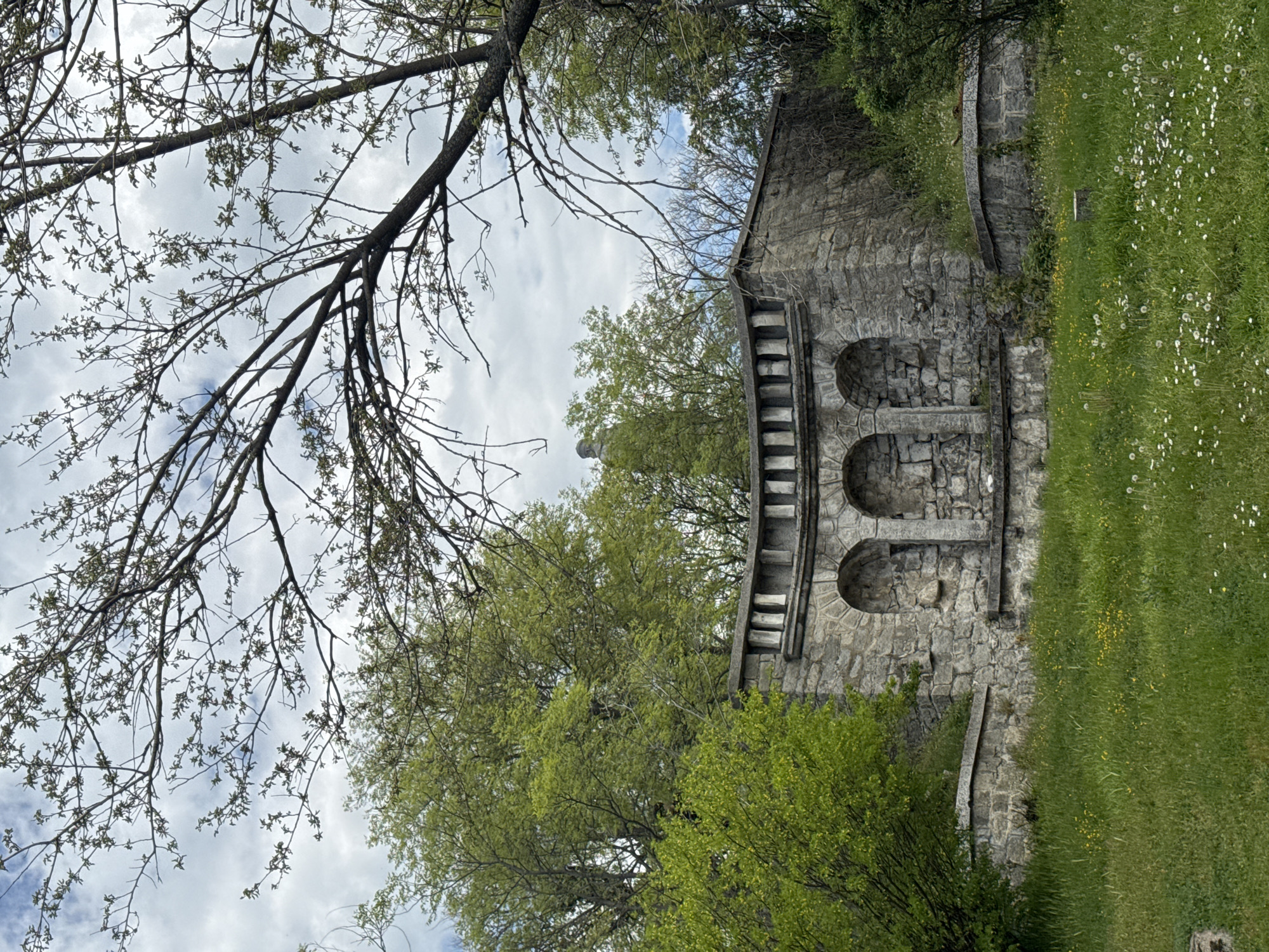 A serene outdoor scene in Plovdiv, Bulgaria, is captured on a partly cloudy spring day. The central feature is an old stone structure, possibly part of a wall or monument, located on the right side of the frame. It is constructed from rough-hewn grey stones and features three stacked arched recesses, with the lower two clearly visible. Along the top edge of this structure, there's a distinctive element resembling a series of stone steps or a decorative balustrade.
To the left and in the foreground, a vibrant green grassy slope is dotted with numerous small white wildflowers, likely dandelions, and some yellow ones, characteristic of springtime. Lush green deciduous trees, with their leaves in full bloom, are visible in the mid-ground and background, partially obscuring the sky behind the stone edifice. In the upper-left quadrant, the intricate, dark branches of a large tree, still mostly bare but with tiny green buds, extend across the sky. The sky shows patches of blue amidst soft white and grey clouds, indicating a pleasant daytime setting. No people or text are visible, and the scene conveys a sense of historical tranquility and natural beauty.