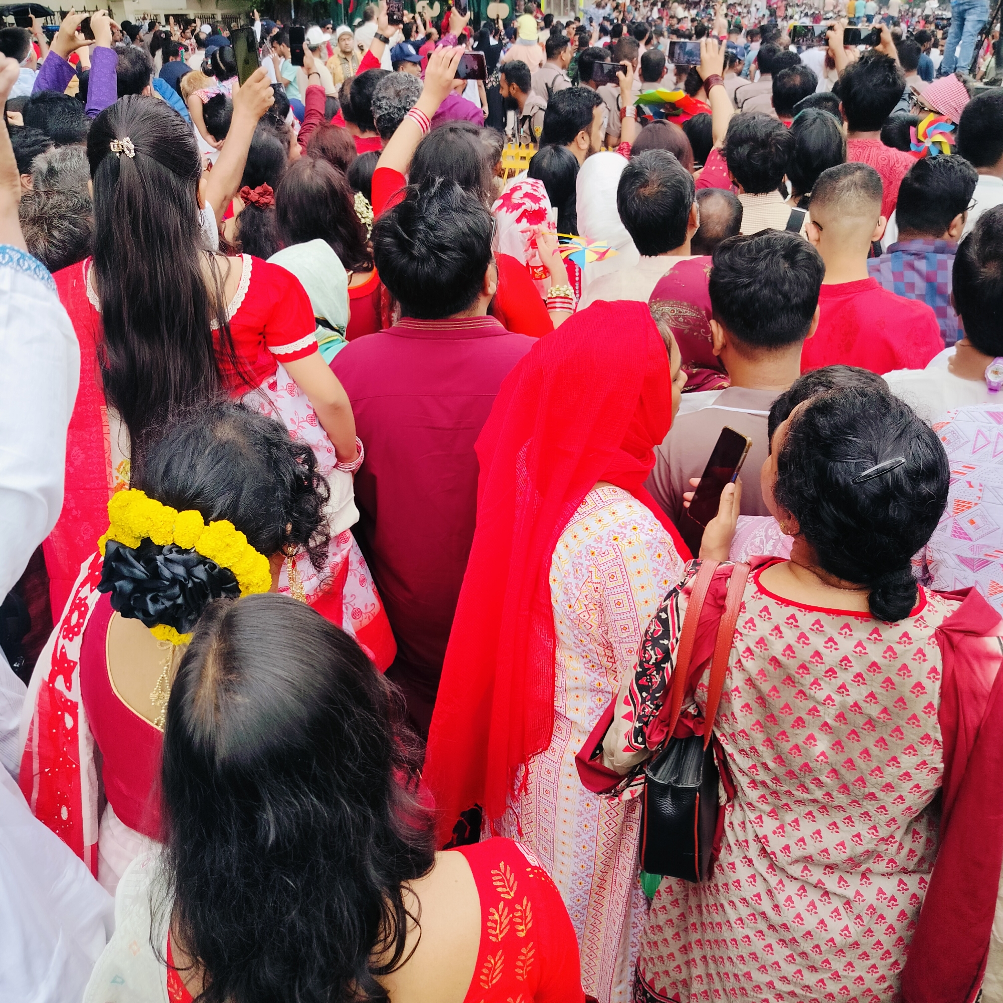 A densely packed crowd fills an urban space, likely a street or public square, in Dhaka, Bangladesh. The majority of people are facing the same direction, suggesting an event or spectacle occurring in front of them. Many individuals hold up their mobile phones, indicating they are taking photos or videos.
Predominantly, the crowd consists of adults, with a variety of hairstyles visible from the rear. Traditional attire is apparent, with several women wearing red and white sarees, and at least one woman is wearing a red hijab. A few are holding red and white items, possibly flags or decorative pieces.
The background is blurred due to the depth of field, but glimpses of buildings and structures indicate an urban setting. The overall atmosphere appears festive and celebratory.