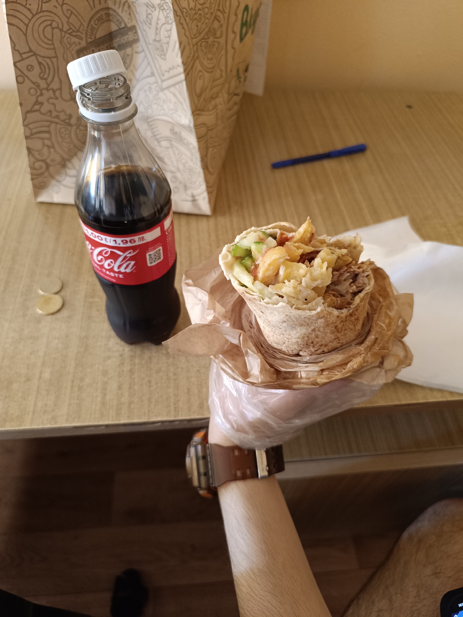 The image depicts a person seated at a light-colored wooden table. The person's left arm is visible, with a G-Shock watch. The person is holding a shawarma in a plastic-gloved hand. On the table is a small glass bottle of Coca-Cola with a white cap, two coins, a pen, a white napkin and a brown paper bag. The price tag on the bottle indicates 1.96 lv (Bulgarian levs). Based on the currency, the location is likely Sofia, Bulgaria. The scene suggests the person is about to eat the shawarma with a drink.