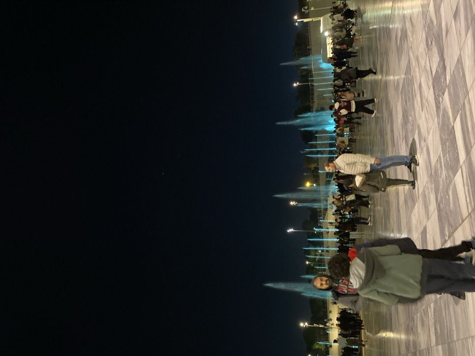 The image captures a vibrant night scene in Kallithea, Greece, centered around illuminated fountains and a gathering of people. The composition is oriented vertically, with a dark, clear night sky occupying the left portion and a brightly lit public square on the right.
In the foreground, two individuals are prominent. A man wearing a light-colored jacket, blue jeans, and sneakers stands with his back to the fountains, looking towards the camera's general direction. His figure, along with other elements, casts a clear reflection on the highly polished or possibly wet, light and dark grey tiled ground. Closer to the viewer, another man with a beard is partially visible, facing slightly towards the camera, alongside a child or smaller person in a green/grey hooded jacket.
Further back, a multitude of people are gathered, forming a crowd that extends along the illuminated area. They appear to be observing several water fountains, which are actively spraying jets of water into the air. These water jets are dramatically lit with a bright blue light, creating a striking visual effect against the dark background. Multiple lampposts or streetlights are also visible, providing additional illumination to the plaza. In the distant background, some indistinct buildings or structures can be discerned. The overall impression is one of a lively public space where people are enjoying an evening display of illuminated water features. No text is visible in the media file.