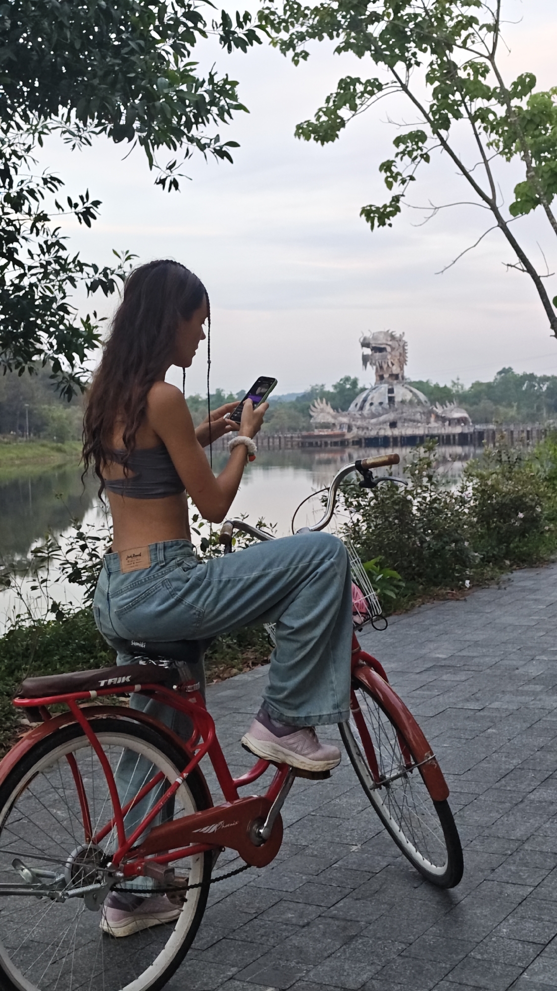 A young woman sits on a red bicycle, holding a cell phone in her hands, near Hồ Thủy Tiên Lake in the Thủy Xuân Ward of Vietnam. The lake reflects an abandoned dragon-shaped waterpark structure on the water's surface. The woman wears a gray top and light blue jeans and purple sneakers. The bicycle, marked with "TRIK" on the seat, rests on a paved pathway bordered by green vegetation. The background shows trees around the lake. The sky is overcast.