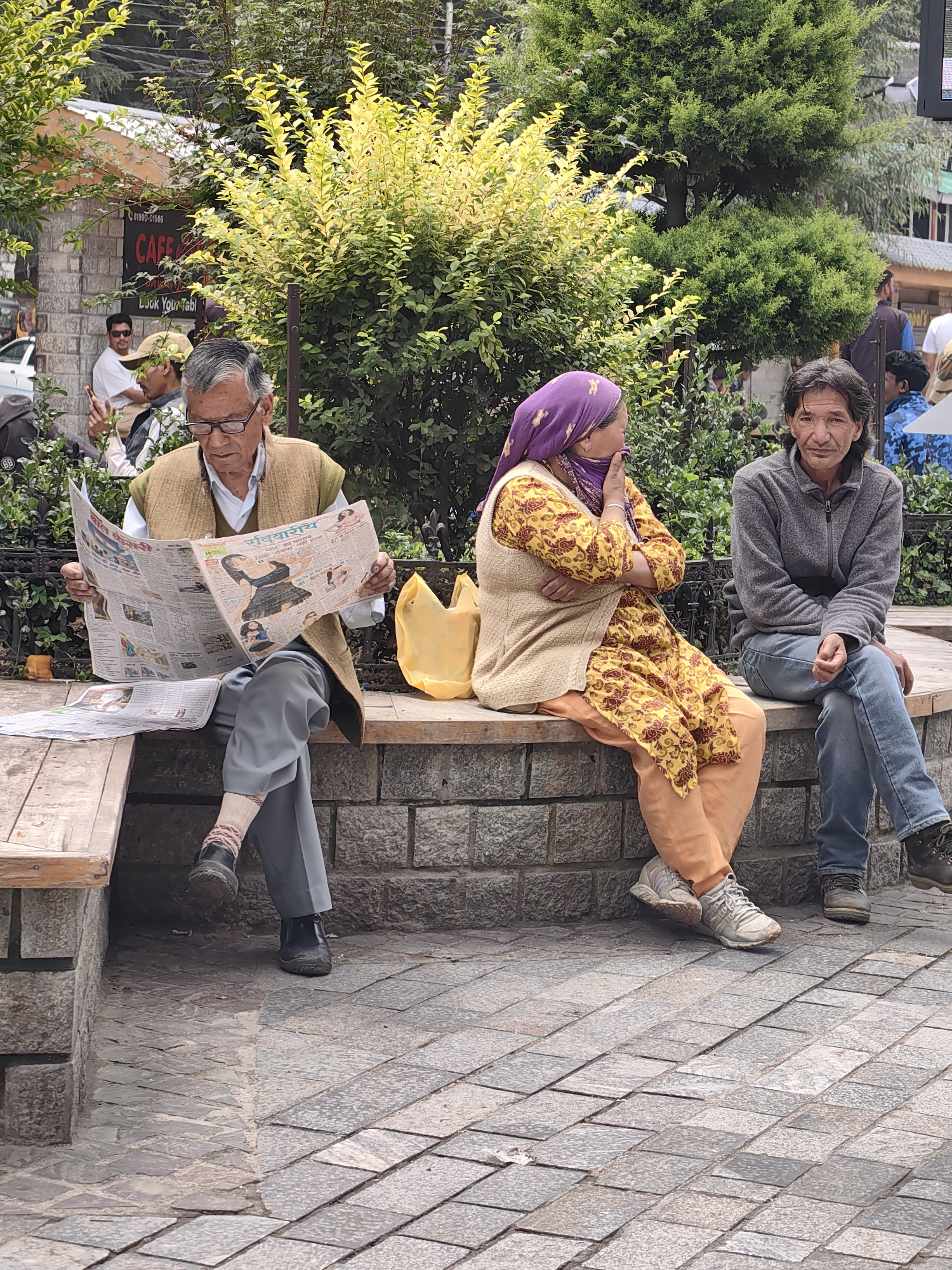 The image captures an outdoor scene in Manali, India, likely a public seating area or a cafe vicinity. Three individuals are seated on a stone bench in front of a backdrop of lush greenery and a brick wall.
On the left, an elderly man wearing glasses and a light brown vest over a white shirt and grey pants is engrossed in reading a newspaper. He is sitting on a wooden bench. To his right, a woman is seated, wearing a purple headscarf, a yellow patterned dress with red accents, and a light beige vest. She is looking away from the camera with her hand near her face. To her right, a man in a grey zip-up jacket and blue jeans is seated, looking directly at the camera. A bright yellow bag is placed on the stone bench between the woman and the elderly man.
The environment suggests a relaxed setting, possibly a tourist spot, with the presence of cafe signage visible in the background. The ground is paved with stone tiles.