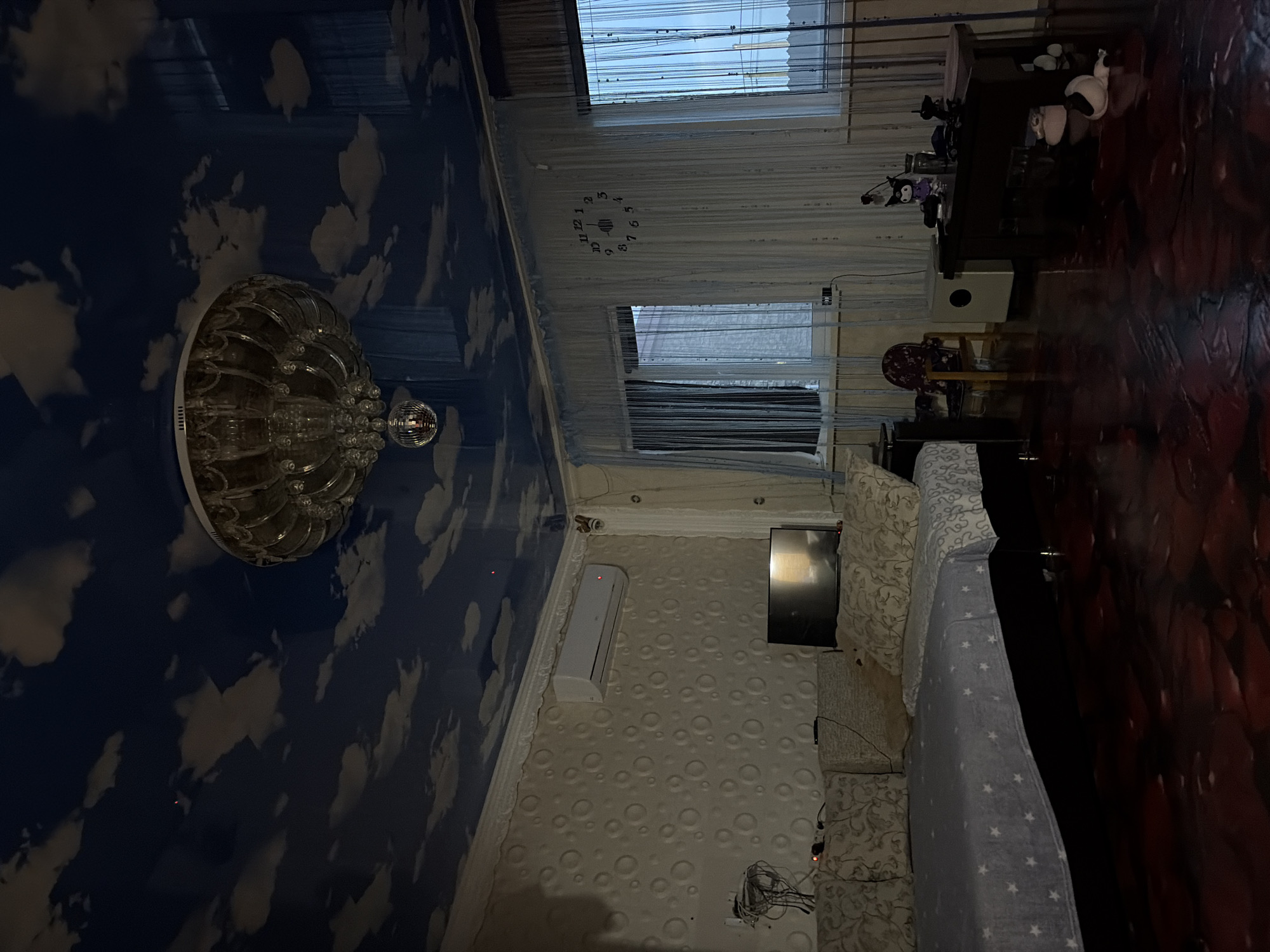 The image depicts a dimly lit living room. The ceiling is a dark blue with a cloudy sky pattern and features a large, ornate chandelier with a disco ball hanging below it. The walls have a textured, circular pattern. On the left side of the room, a sofa is covered with a light blue throw blanket with white stars. An air conditioning unit is mounted on the wall above the sofa. In the center of the room, a television is placed on a surface. To the right, there are several windows covered by sheer curtains. A large wall clock with black numbers is visible on the wall between two windows. In the foreground, the floor is dark and appears to have a glossy, reflective surface with a reddish-brown hue. On a dark wooden table to the right, various objects are placed, including figurines and what appears to be a jar. A small chair is also visible near the windows. The overall impression is of a residential interior.