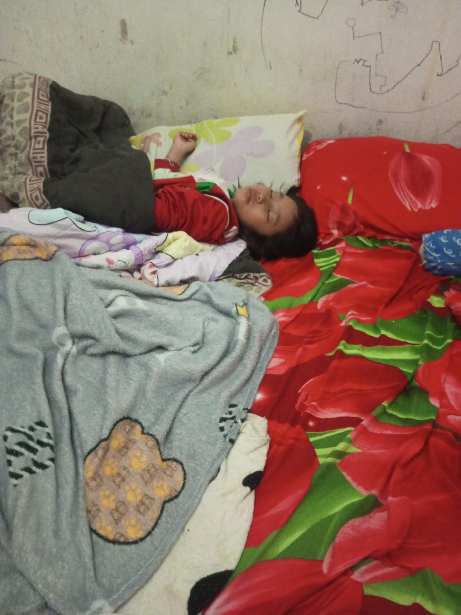A young child sleeps peacefully on a bed. The child is lying on their back, with their head resting on a pillow. They are covered by a grey blanket with a bear design. The bed is adorned with floral-patterned sheets in red and green. In the background, a wall is visible, decorated with a child's drawing. This scene is likely in a residential setting within Andongsili, Indonesia.