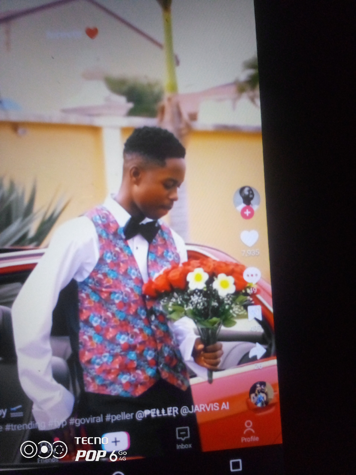 A young man, dressed in a white shirt, a black bow tie, and a colorful vest with a floral pattern, stands outdoors and holds a bouquet of red roses and white flowers. He is positioned in front of a red car. The background shows a building with yellow walls and some greenery. On the right side of the image, social media interface elements are visible, including a profile picture, a heart icon with a number "7,935" below it, and comment icons. The bottom of the image displays text that reads "TECNO POP 6 Go 4G" along with other interface elements that suggest a smartphone screen. The scene appears to be in an urban setting, possibly the city of Owerri, Nigeria, given the brand of the phone and the general aesthetic.