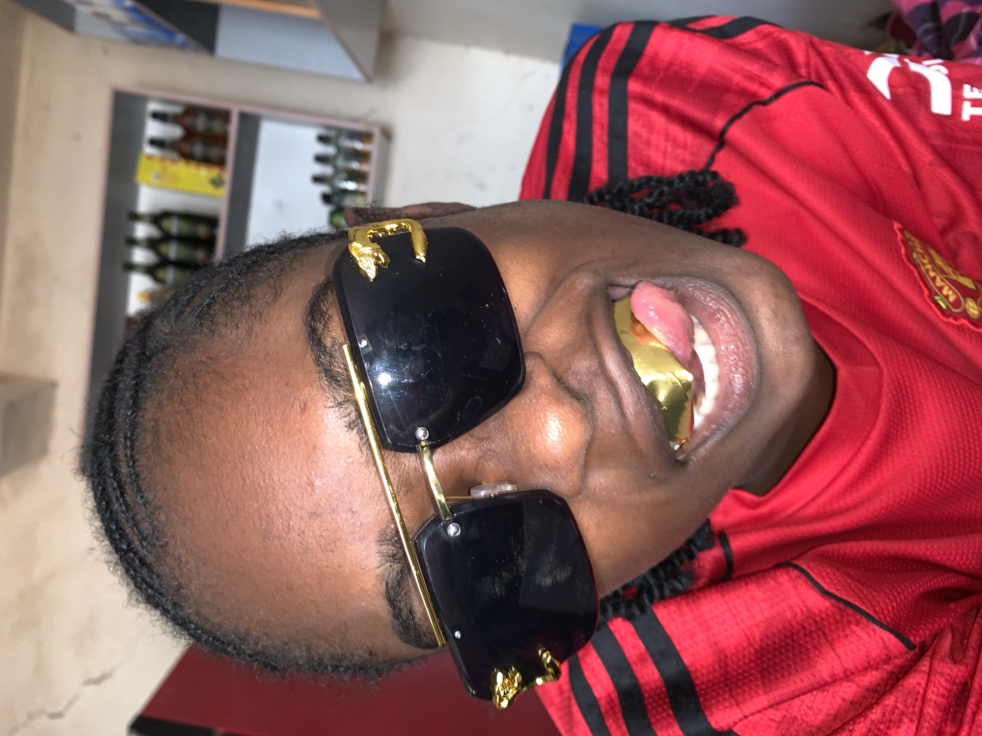 A close-up portrait, oriented sideways, captures a person with dark skin, wearing a red athletic jersey and large black sunglasses with gold frames. The subject's head is tilted, and their mouth is open, prominently displaying a shiny gold grill covering their upper teeth, with their tongue slightly visible.
The red jersey features black stripes on the shoulders and a partially visible logo on the chest, which appears to be a Manchester United crest along with the text "MANCH", indicating a Manchester United football club shirt. The sunglasses have decorative gold embellishments on their upper corners. The person's hair is styled in short braids.
The background, largely out of focus, shows an indoor setting with shelves stocked with numerous bottles, likely alcoholic beverages, suggesting a store, bar, or similar establishment. The lighting is dim, indicating an indoor environment. The pose and accessories convey a confident and stylish demeanor. The image is contextualized to be in New Stadium, Nigeria.