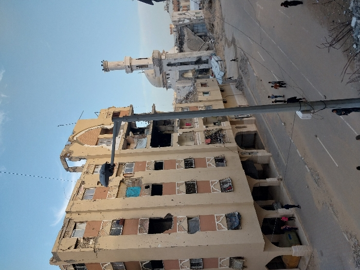 The image depicts a severely damaged urban street scene in Khan Yunis, Palestinian Territory, under a clear blue sky. A multi-story beige building, likely an apartment block, dominates the left side, exhibiting extensive structural damage including numerous impact marks, shattered windowpanes, exposed interiors, and a partial collapse of its upper facade. Torn curtains and a piece of blue fabric are visible hanging from some windows. To the right, a mosque featuring a white dome and a tall, slender minaret stands amidst significant widespread destruction, with multiple adjacent structures reduced to large piles of concrete rubble and twisted rebar. The paved street, covered in dust and scattered debris, features a prominent grey street light pole in the mid-ground. Several groups of civilians, including adults and children, are visible on the road and under the damaged building's arched ground-floor section, either walking or standing amidst the devastated environment.