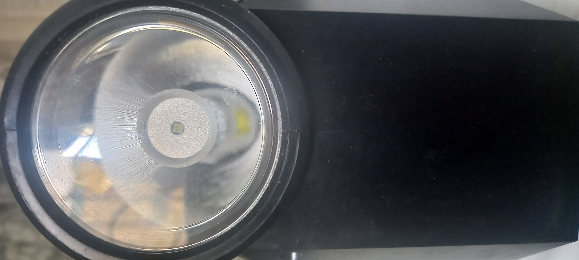 The image shows a close-up of a black flashlight. The lens of the flashlight appears to have moisture or condensation inside. The environment is not discernible enough to determine location.
