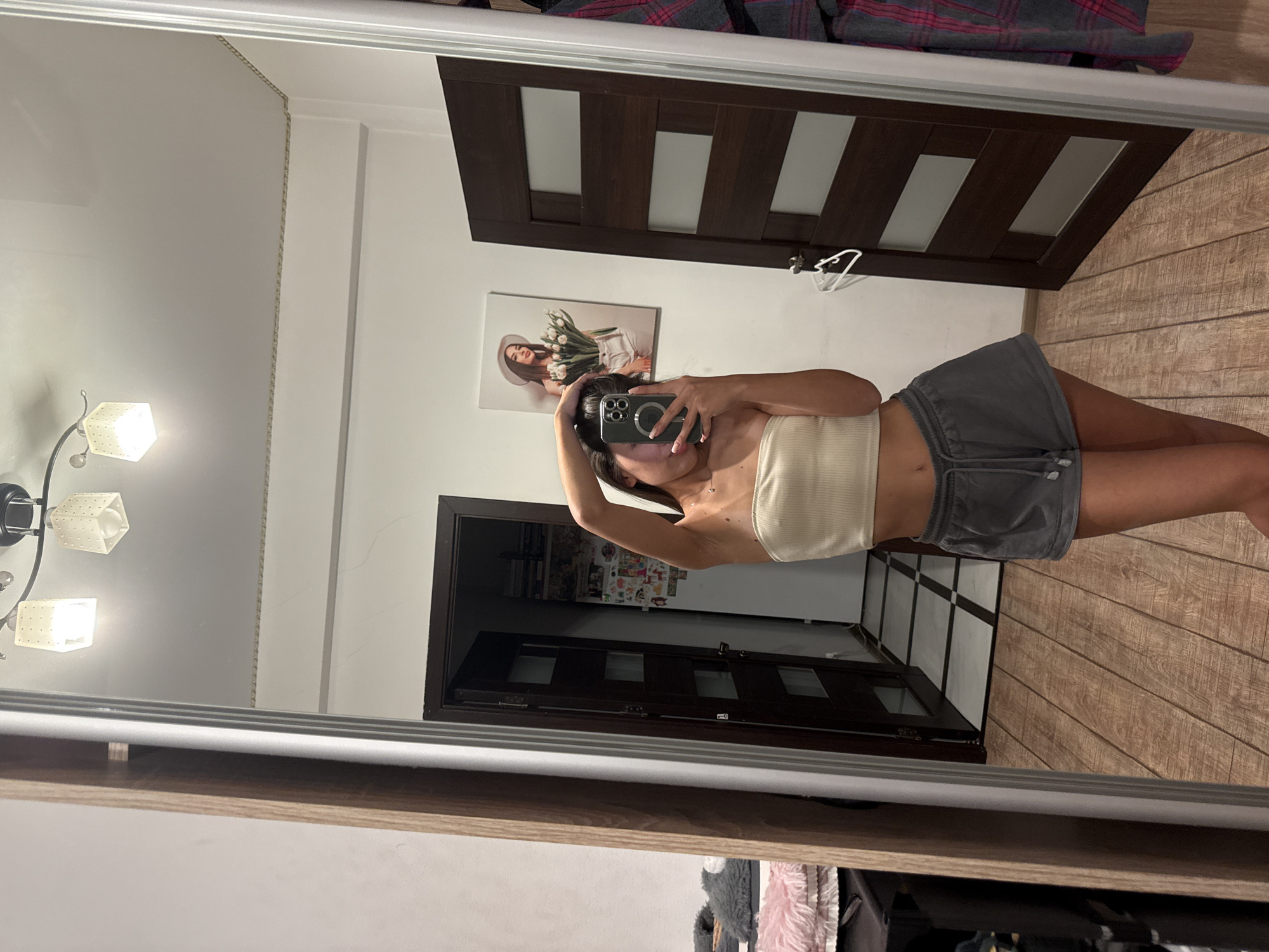 A young woman with dark hair is taking a mirror selfie in a room. She is wearing a light-colored strapless top and gray shorts. Her right arm is raised, and she is holding her hair up with her left hand as she holds a smartphone in her right hand to capture the image. In the background, a large mirror reflects the room, showing two closed dark wooden doors with glass panels, one with a white hanger clipped to it. To the left of the doors, a framed picture of a woman wearing a hat and holding a bouquet of tulips hangs on the wall. A tiled floor is visible beyond the open doorway. The room appears to be a domestic setting.