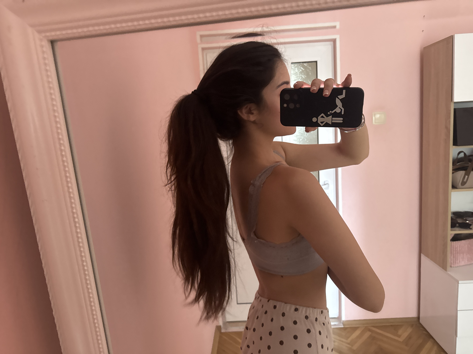A young woman is depicted taking a mirror selfie in an indoor setting in Plovdiv, Bulgaria. She stands with her back partially turned towards the camera, looking at her reflection in a large mirror with a decorative white frame. Her long, dark hair is styled in a high ponytail. She is wearing a light grey, ribbed bralette or sports bra and light-colored shorts adorned with dark polka dots.
She holds a black smartphone horizontally with both hands, its screen obscured by her hands. The phone case features a white graphic showing stylized stick figures: a female figure standing, followed by an equal sign, and a male figure doing a handstand above her, with another male figure appearing to tumble to the right. Her fingernails have a neat French manicure.
The room features soft pink walls and a light wooden floor with a herringbone pattern. Visible in the reflection are part of a white door frame and a section of a white and light wood cabinet or wardrobe with shelving units containing various items. The presence of natural light suggests the image was taken during the daytime. The subject's facial expression is not discernible as her face is mostly hidden behind the phone.
