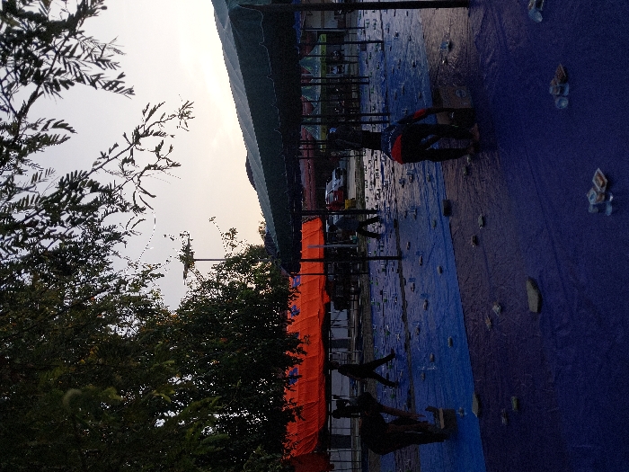 The scene depicts an outdoor area in Lhokseumawe, Indonesia, at either dawn or dusk, indicated by the soft, pale light. The ground in the foreground is covered with large blue tarpaulins, sectioned into a darker blue area closer to the viewer and a lighter blue area further back. Numerous small, clear plastic cups, small rectangular packaged items, and flat, dark, irregularly shaped objects (possibly stones or weights) are scattered across these tarpaulins.
Temporary tent structures are visible, supported by slender, dark vertical poles. Some tents feature orange canopies, while others have dark green canopies. In the upper left and central left, green foliage from trees and branches extends into the frame. A light pole with a solar panel and lamp head is positioned in the upper mid-left background. Beyond the tarp-covered area, a paved surface with several parked vehicles, including a white and a red vehicle, is visible.
At least five individuals, appearing to be adult males, are present. Two individuals in the foreground are bent over, interacting with the items on the blue tarpaulins. The person on the left wears dark clothing, while the person on the right wears a dark top with red/orange accents. Two additional individuals are walking in the mid-ground: one in dark attire moving right, and another in lighter attire moving left. A fifth individual is faintly visible further in the background near the vehicles. The actions of the individuals suggest activity related to the setup or cleanup of an event or market.