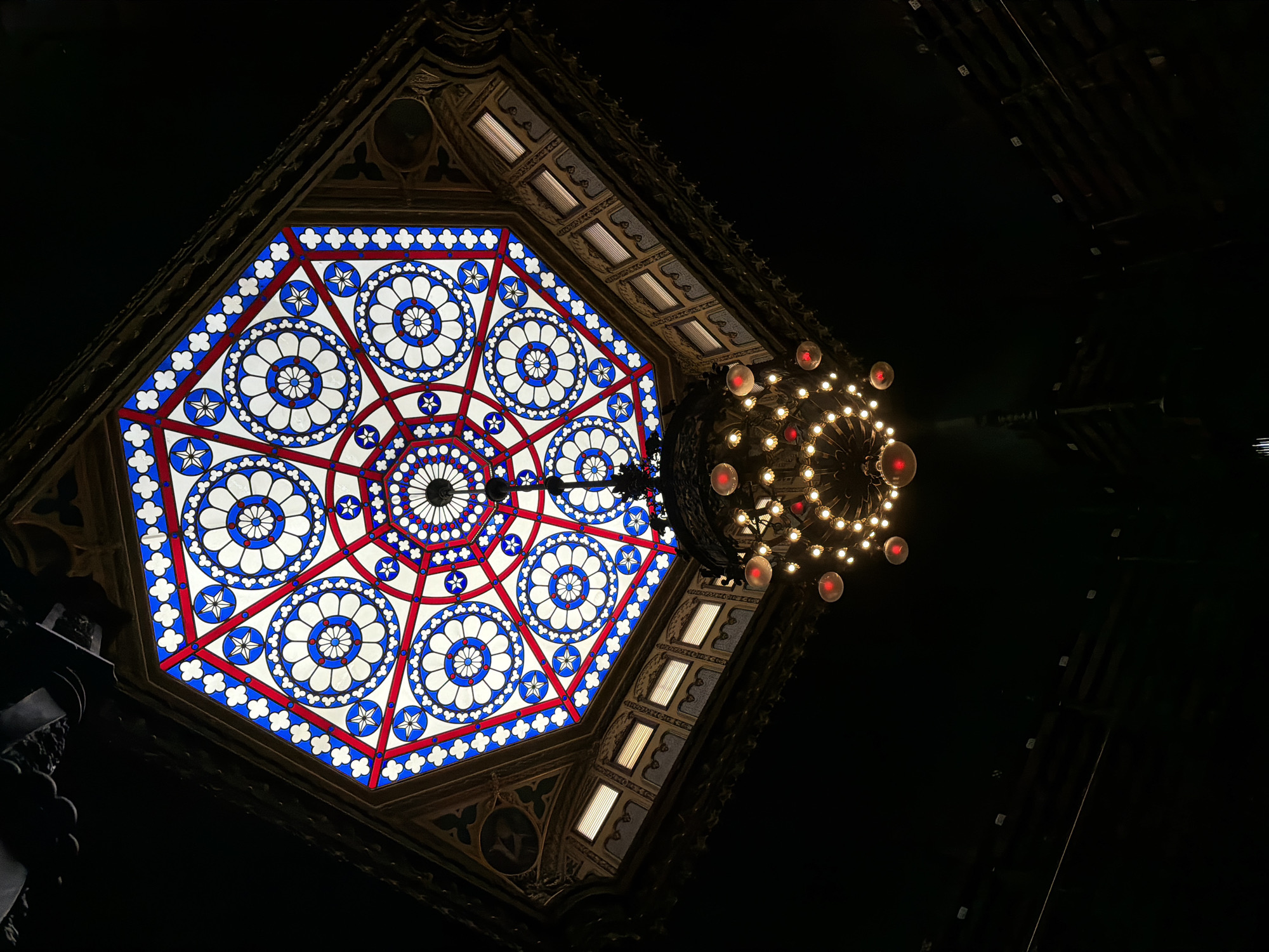 This image shows a view looking up at a stained-glass octagonal skylight and a chandelier in what appears to be a grand interior space. The skylight features a geometric pattern of red, white, and blue glass, with circular and star motifs. Below the skylight, a multi-tiered chandelier with numerous lights illuminates the space. The surroundings are dark, suggesting the photo was taken at night or in a dimly lit area. The architectural details of the ceiling and upper walls are visible, with ornate carvings and what appear to be small windows. The overall impression is one of ornate historical architecture.
The image is of the interior of the National Library of Brazil (Biblioteca Nacional) in Rio de Janeiro, Brazil. Specifically, it shows the skylight and chandelier in the main hall, known as the "Salão Principal" or "Salão de Leitura" (Main Hall or Reading Room). The library itself is a significant historical landmark located in the Cinelândia square in downtown Rio de Janeiro.