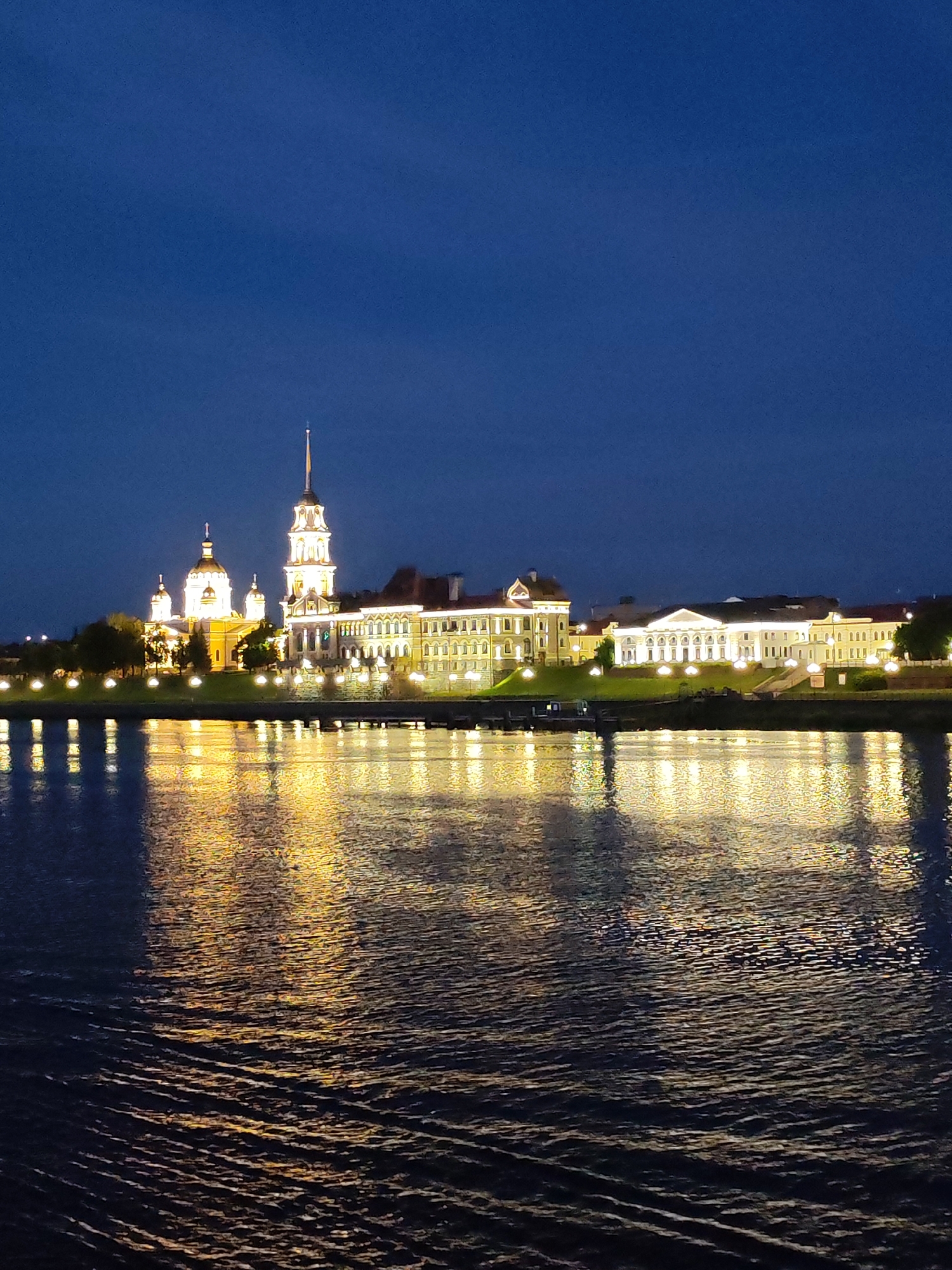 The image depicts a nighttime view of a waterfront with illuminated buildings and their reflections on the water's surface. On the left, a church with multiple domes, one large and central, is lit, with smaller domed structures nearby. Adjacent to the church, a tall bell tower with a spire rises, also illuminated. To the right of the bell tower, a long, multi-story building with many windows is lit from within, its facade a pale yellow. Further to the right, another large, light-colored building with a classical facade and columns is visible, also illuminated. Trees line the embankment between the buildings. The water in the foreground is dark, with wavy ripples reflecting the yellow and white lights from the buildings, creating a shimmering effect. The sky above is a deep, dark blue, indicating nighttime. The location is identified as Bol'shie Mkhi, Russia.