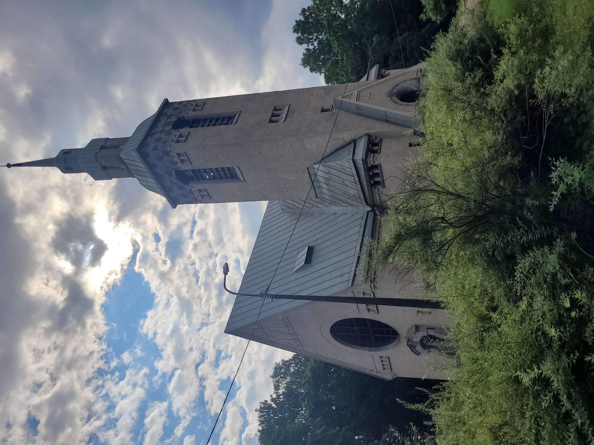 This is a full-frame, low-angle, eye-level shot of a church in Zelenogorsk, Russia. The church, constructed of light-colored stucco, features a tall, slender tower topped with a spire and a steeply pitched metal roof. The tower has several rectangular windows and decorative stone detailing at its base. A large, circular stained-glass window is visible on the side of the main church structure, which has a similarly pitched metal roof. The building is partially obscured by dense green foliage in the foreground, which appears to be bushes and trees. A street lamp on a dark pole stands to the left of the church. The sky above is dramatic, with a mix of dark, heavy clouds and bright, sunlit patches. The overall impression is of a somewhat overcast day. There are no people or animals visible in the image.