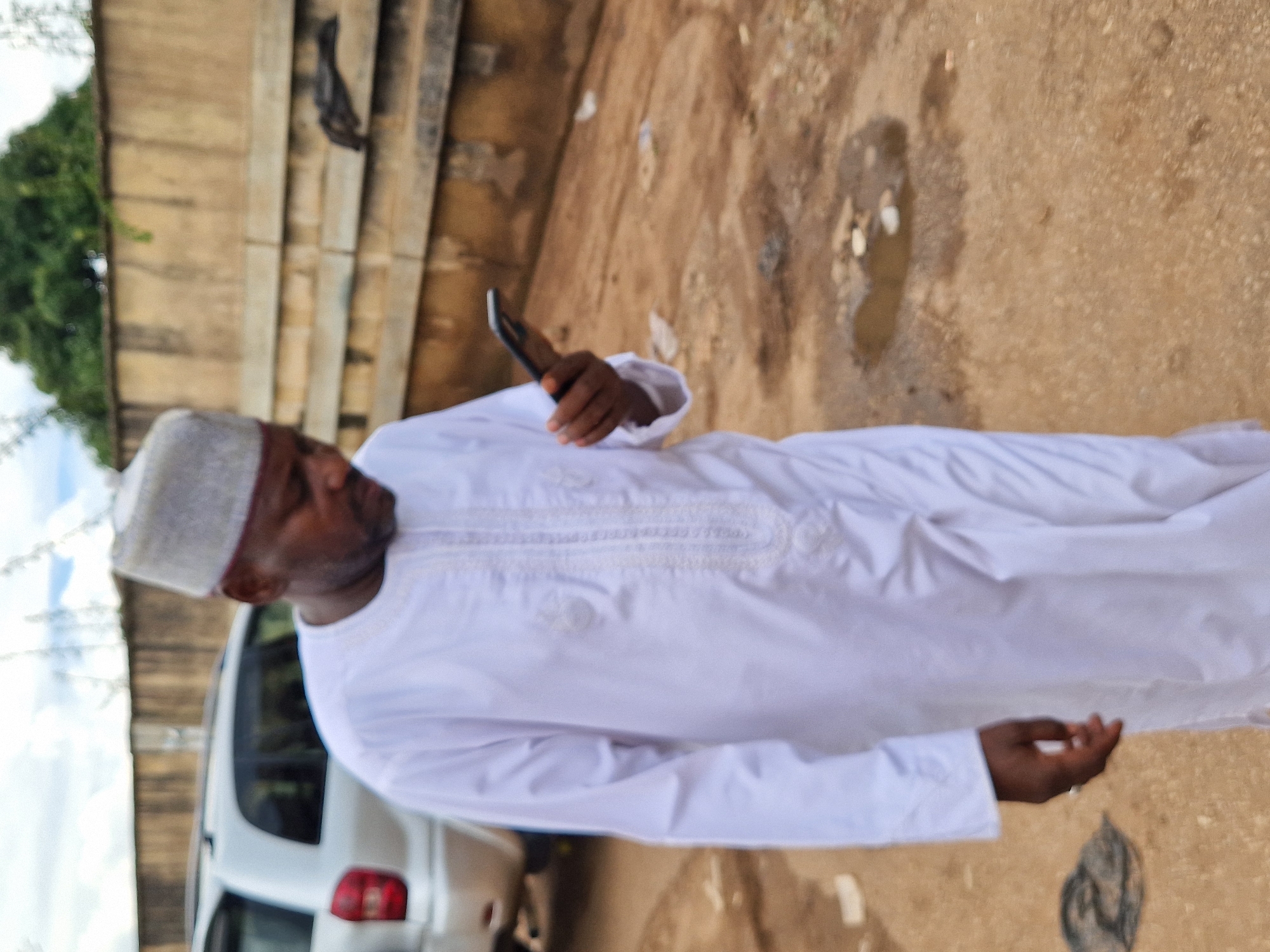 A man in a white traditional outfit and a grey cap is walking and looking at his smartphone. The man is of African descent and appears to be in his 40s or 50s. He is walking on a dirt path in what appears to be an outdoor area, possibly a compound or a less developed part of a city. The background shows a beige-colored concrete wall, a silver SUV, and some greenery. There are puddles of water on the ground, suggesting it might have rained recently. The overall scene is simple and everyday, with no indication of any particular event or activity. The location is identified as Jos, Nigeria.