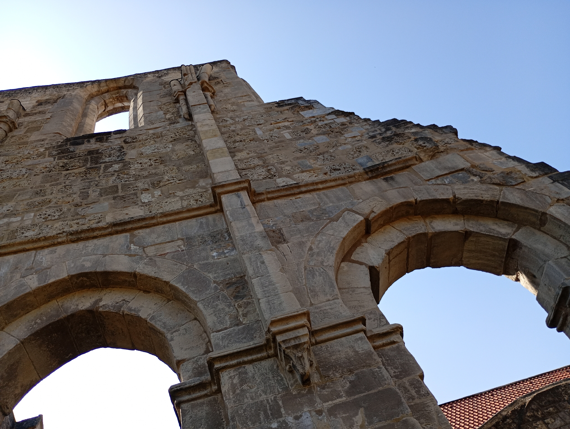 The image shows a low-angle view of the ruined facade of the Walkenried Abbey, located in Walkenried, Germany. The stone structure, composed of weathered, rough-hewn blocks, features two large arched openings. The arch on the left is partially obscured by the perspective, while the arch on the right is more fully visible, framing a clear blue sky. Above the arches, the remaining wall rises, displaying intricate stonework and architectural details, including what appear to be corbels or decorative elements. The top of the wall is uneven, indicating its ruined state, with some sections showing jagged edges against the bright sky. In the lower right corner, a portion of a red-tiled roof is visible, suggesting the presence of a more modern structure adjacent to the abbey ruins. The lighting suggests it is daytime, with sunlight illuminating the stone surfaces and creating a contrast with the shadows within the arches and on the textured stone.