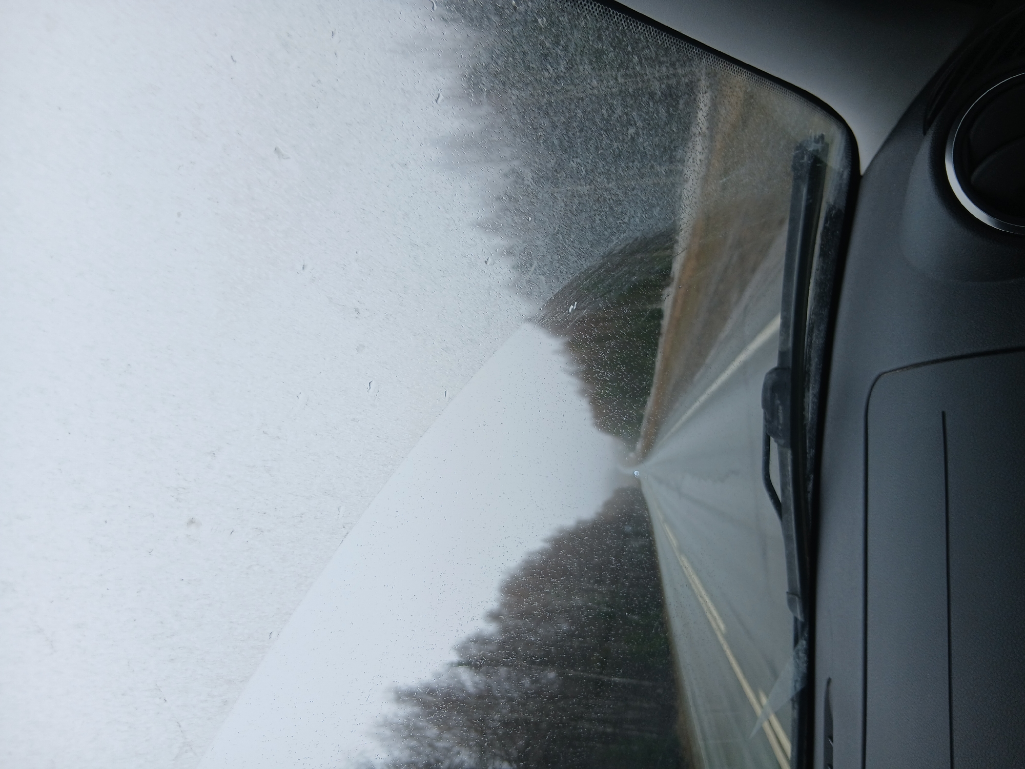 The image depicts a view from inside a vehicle, looking through the windshield. It is raining, and the windshield wipers are active. A road stretches ahead, flanked by trees on both sides. The environment appears overcast and wet. The dashboard of the vehicle is visible on the right side of the frame, including an air vent. The location is not discernible from the image. There are no visible people.