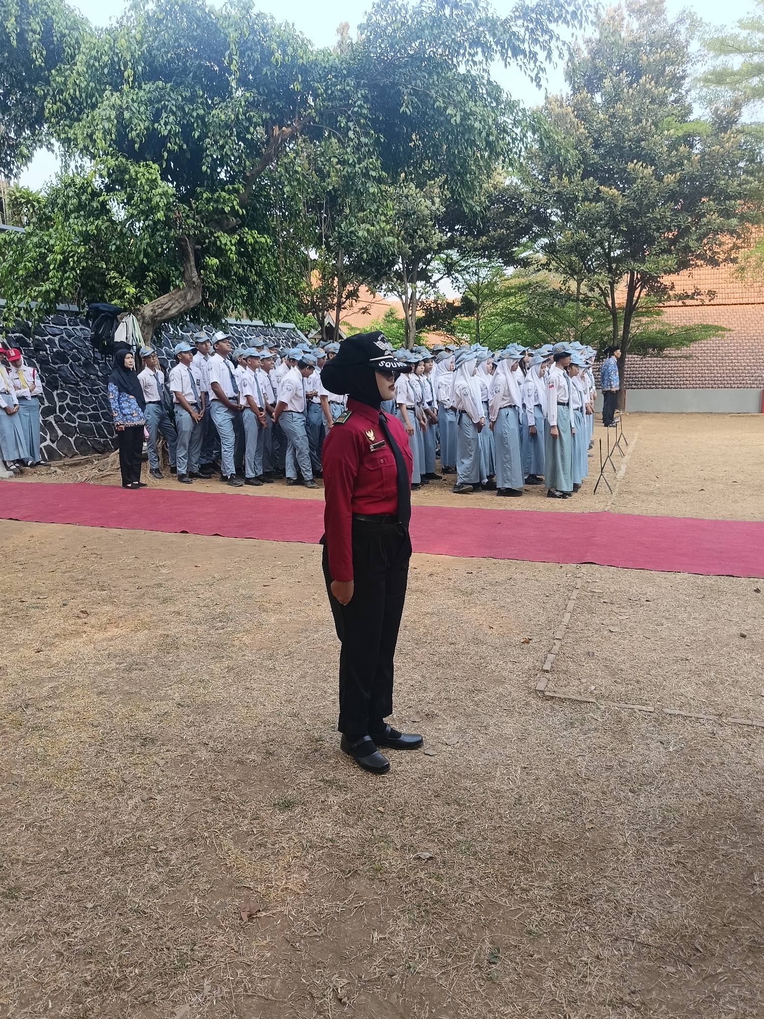 In Pengkol, Indonesia, a group of students, likely from a school given their uniforms, are lined up outdoors on a grassy area. A prominent red carpet stretches across the ground in front of them. A young woman in a distinctive red uniform and black head covering stands at attention in the foreground, facing away from the camera. Behind her, a line of students in white shirts, light blue pants or skirts, and white hats are organized. To the left, a woman in a blue and white patterned top and black pants stands with a group of students. In the background, large trees with dense green foliage are visible, along with a brick building and a stone wall. The overall atmosphere appears to be that of an organized event or ceremony.