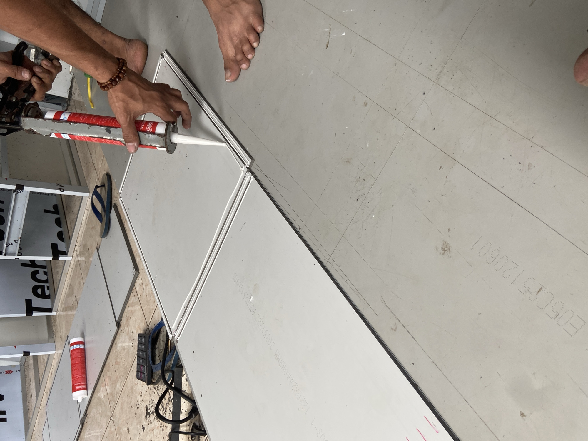 The image depicts a close-up view of a person's hands applying sealant from a caulking gun onto the seam between two large, light gray panels laid flat on a floor. The panels appear to be part of a construction or installation project. The floor itself is also tiled in a similar light gray color, with visible grout lines forming a grid pattern. In the background, partially visible structures with the text "Tech" suggest a commercial or industrial setting. There is a red and white tube of sealant on another panel to the left, and a black calculator and electrical cord nearby. The person's bare feet are visible on the floor, one in the foreground and another towards the right of the image. The lighting is moderately bright, casting some shadows. The scene appears to be taking place indoors, possibly a workshop or a completed section of a building. There is no identifiable information in the image to indicate the location in the city of Plosokandang, Indonesia.