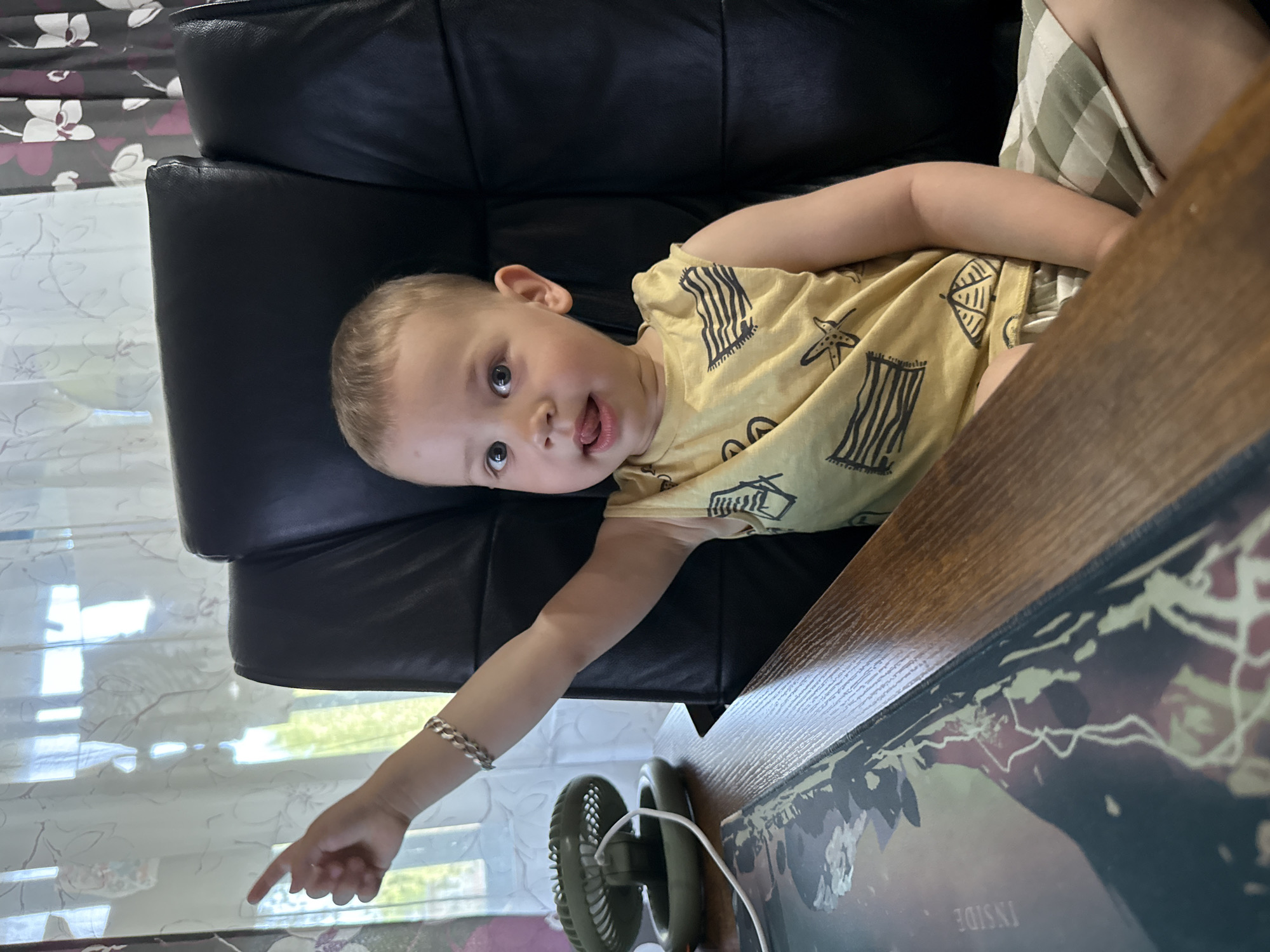 A young child is lying on a black leather chair next to a wooden desk. The child is wearing a yellow tank top with black designs. The child is pointing with their right hand towards a small green fan on the desk. A silver bracelet is on the child's right wrist. The desk has a book or folder on it. A window with floral curtains is visible in the background. The location is Rusanovo, Russia.