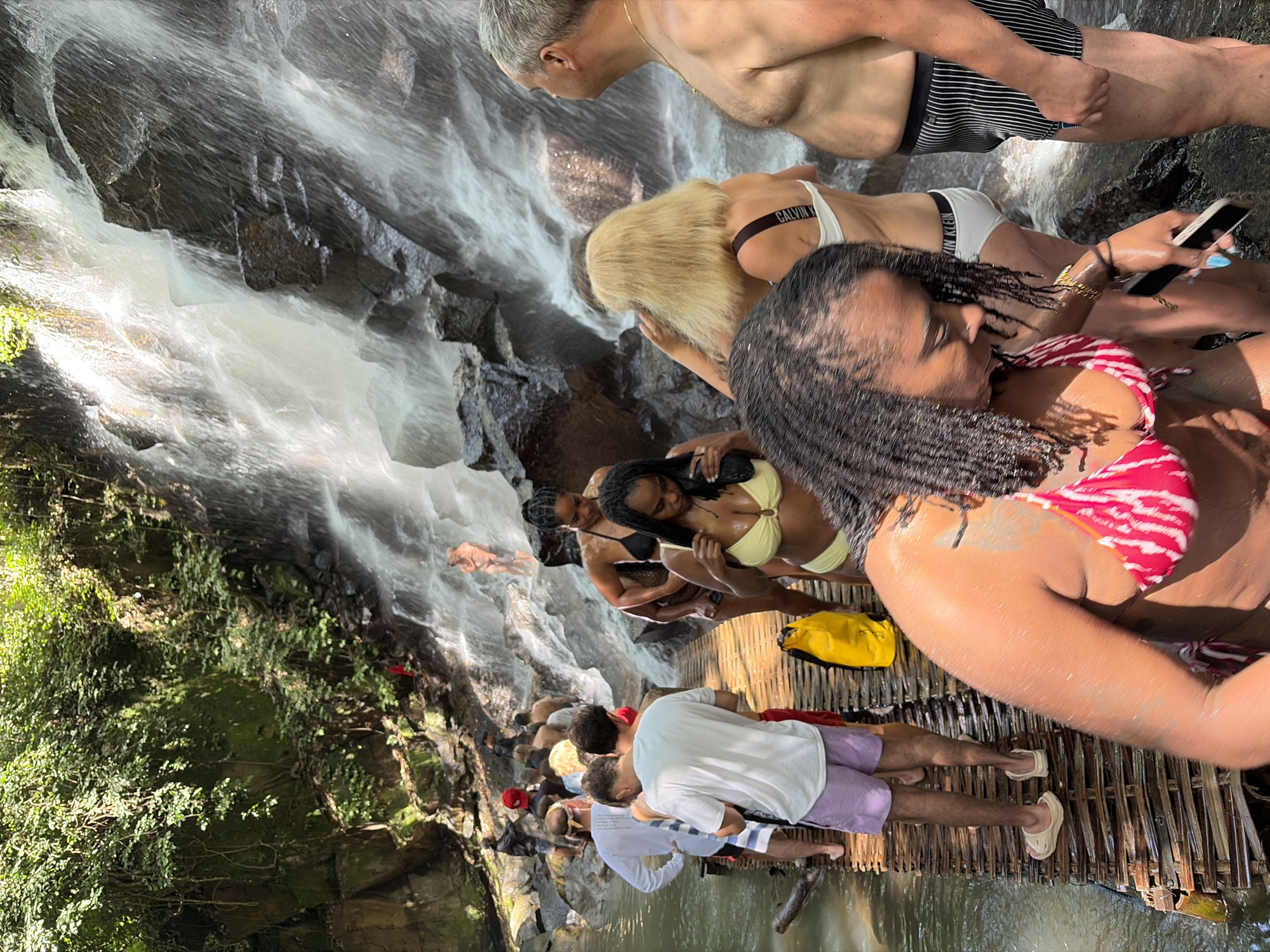 A group of people are gathered on a wooden walkway in front of a waterfall. The waterfall cascades down a rocky cliff face covered in lush green vegetation. The people are dressed in swimwear, and some appear to be posing for photos or waiting to enter the water. The walkway is made of interwoven bamboo or wood planks. The water appears to be clear and cool. The scene suggests a popular tourist destination known for its natural beauty.