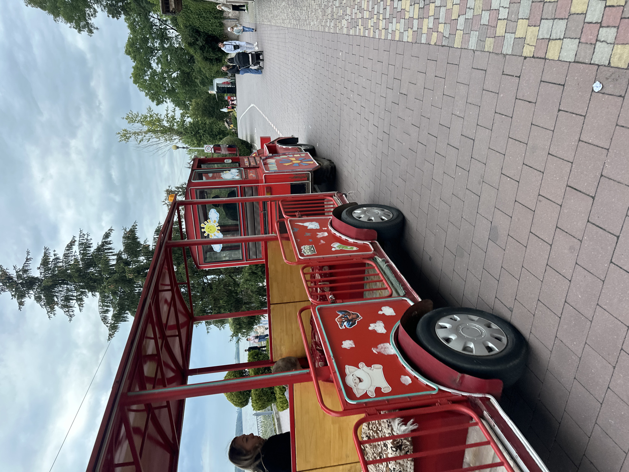 In Ternopil, Ukraine, a vibrant red tourist train is parked on a paved walkway. The train consists of multiple open-air carriages adorned with colorful cartoon decals, including a sun, clouds, and animal figures. A woman with blonde hair is seated in the first carriage, facing away from the camera. Another person, possibly a child, is partially visible inside the same carriage, peeking over the wooden interior. Further down the walkway, a family walks with a stroller, and other individuals are seen in the background, suggesting a public recreational area near a body of water, likely Ternopil Lake. The sky is overcast with clouds, and lush green trees line the background. The walkway is constructed with interlocking pavers, with a section featuring a colorful mosaic pattern. The overall scene depicts a leisurely day in a Ukrainian city.