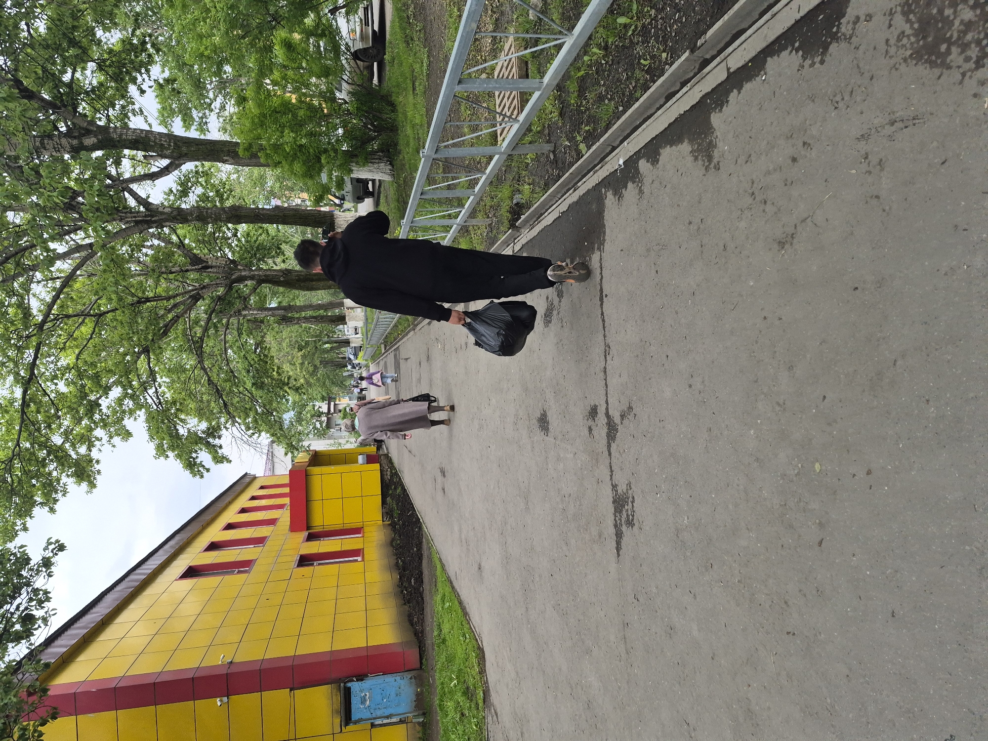 The image depicts a paved walkway in Uglegorsk, Russia, adjacent to a bright yellow building with red trim. The walkway is lined by a metal fence and trees. Several people are visible walking away from the camera. A man in a black hoodie and pants is carrying a black plastic bag in his right hand and appears to be talking on his phone. Further ahead, an elderly person in a long grey coat walks with a handbag. A child in a pink outfit is seen behind them, and other figures are further in the distance. The environment suggests a residential or public area with typical urban landscaping.