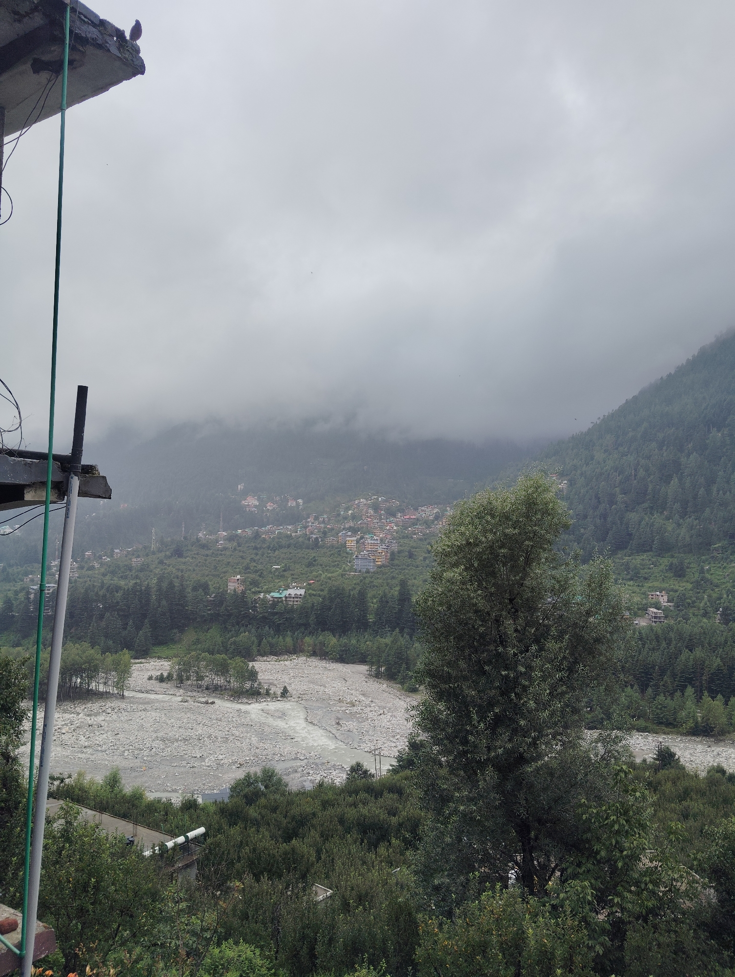 The image depicts a landscape in the city of Vashisht, India, characterized by a cloudy sky over a verdant, mountainous terrain. In the foreground, a concrete structure juts out from the left edge, with a bird perched on its corner. A green pipe and some wires are also visible attached to this structure.
Dominating the midground is a wide, pebble-strewn riverbed, with a gentle stream of water flowing through it. The banks of the river are lined with trees. Beyond the river, the landscape rises into a densely forested hillside, dotted with numerous buildings of varying sizes and architectural styles, indicative of a settlement. These buildings are mostly clustered together, suggesting a town or village nestled into the hillside.
Further into the background, the mountains are shrouded in thick clouds and mist, creating a soft, diffused light across the scene. The overall impression is one of a somewhat overcast day in a hilly, populated region.