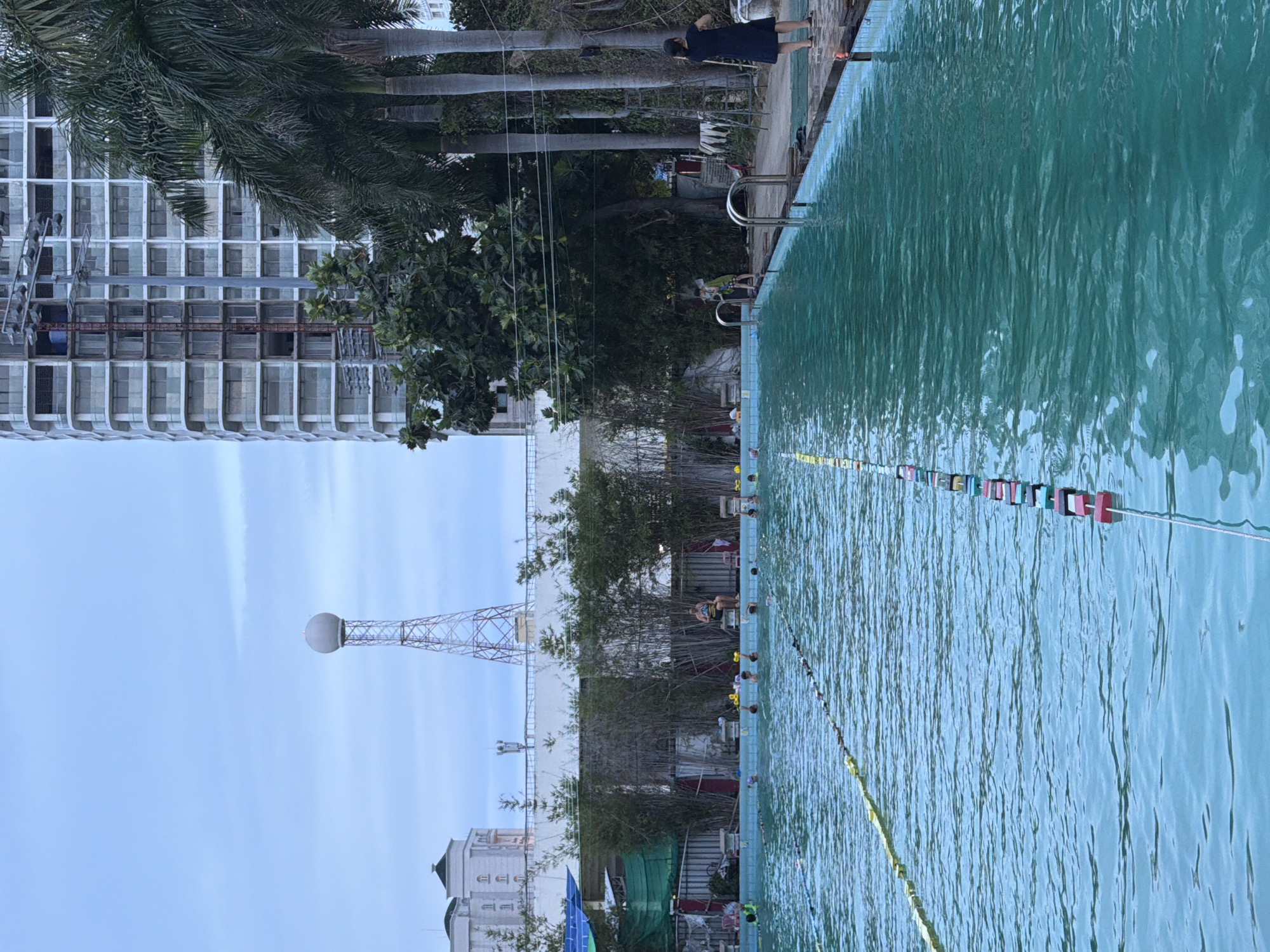 The image shows a swimming pool in Nha Trang Ward, Vietnam. The pool is long and narrow, with lane markers visible in the water. Several people are swimming or wading in the pool. In the background, there are tall buildings, palm trees, and a radio tower. The sky is overcast.