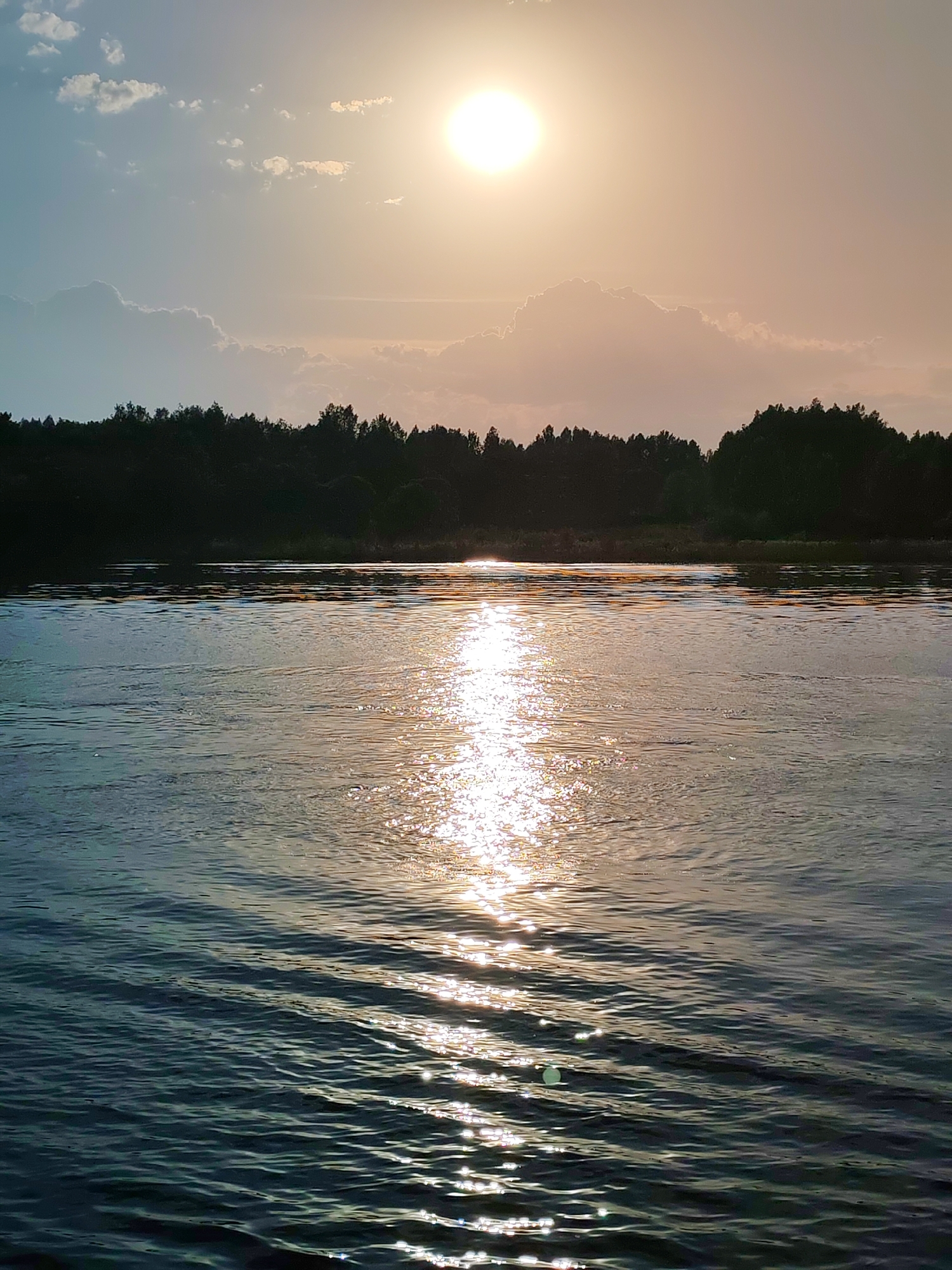 The image captures a serene scene at an unspecified location in Novye Shatrishchi, Russia. The primary subject is a body of water, likely a river or lake, with gentle waves reflecting the bright sunlight. The sun is positioned high in the sky, obscured slightly by thin clouds, casting a golden glow over the scene. A dense line of trees is visible in the background, silhouetted against the horizon. The sky transitions from a light blue on the left to a warm orange-yellow near the sun, indicating either sunrise or sunset. There are no people or objects visible in the immediate foreground. The environment appears natural and undisturbed.
