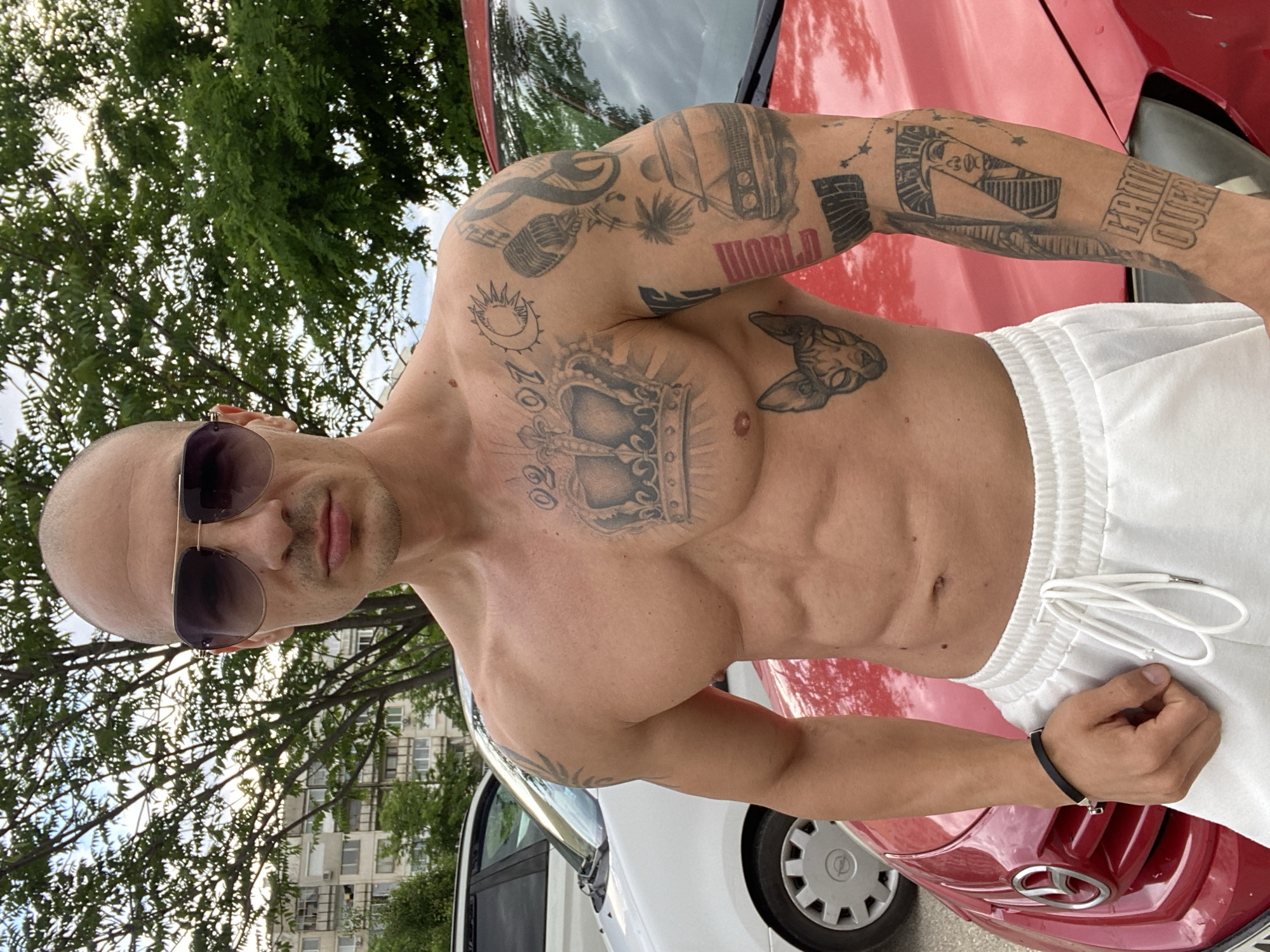 A muscular man is captured in a shirtless, outdoor daytime portrait. He is bald, has light facial stubble, and wears dark sunglasses, looking directly at the camera with a neutral expression. He is wearing white athletic shorts and a black bracelet on his left wrist.
His torso and arms are heavily tattooed. Prominently on his chest is a large crown design, with the numbers "02" (or "ZO") above it, and a sun-like symbol to the side. Tattoos on his right arm (viewer's left) include a boombox, a microphone, a palm tree, and the word "WORLD" in red text, alongside abstract patterns. His left arm (viewer's right) features an Egyptian-style head, the words "FAME QUEEN," and starry/constellation patterns.
The background shows green foliage of trees with sunlight filtering through, indicating a bright, sunny day. Beyond the trees, multi-story residential buildings are visible, suggesting an urban or suburban setting. A red car, possibly a Fiat, is parked to the man's right, with its hood and windshield reflecting the surroundings. Part of a white car, possibly an Opel due to its hubcap design, is visible to his left. The photo is taken in Pleven, Bulgaria.