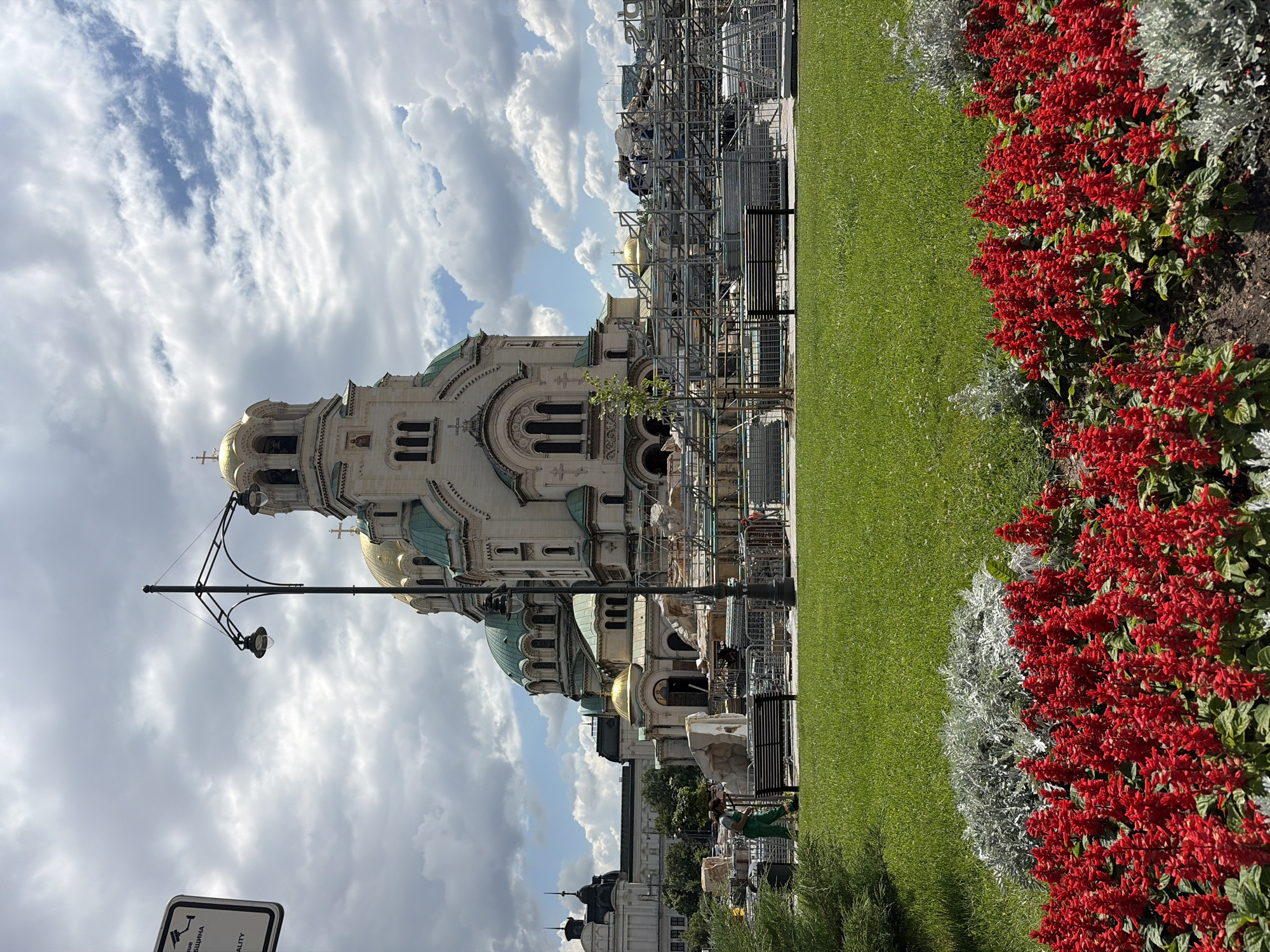 This image captures the exterior of the Alexander Nevsky Cathedral in Sofia, Bulgaria, under a cloudy sky. In the foreground, a manicured lawn is bordered by vibrant red flowers and silvery-grey foliage. A black park bench is visible on the lawn, and a lone figure in green overalls is seen tending to the grounds near the bench. Behind the lawn, scaffolding and construction materials suggest ongoing renovation or maintenance work on the cathedral and its adjacent structures. The cathedral itself, with its iconic golden domes and green roofs, is the central architectural element, with other buildings visible in the background. A lamppost stands in the middle ground, and a sign with text in Cyrillic script is partially visible on the left. The overall impression is one of a significant landmark undergoing some form of upkeep, with elements of urban landscaping in the foreground.
