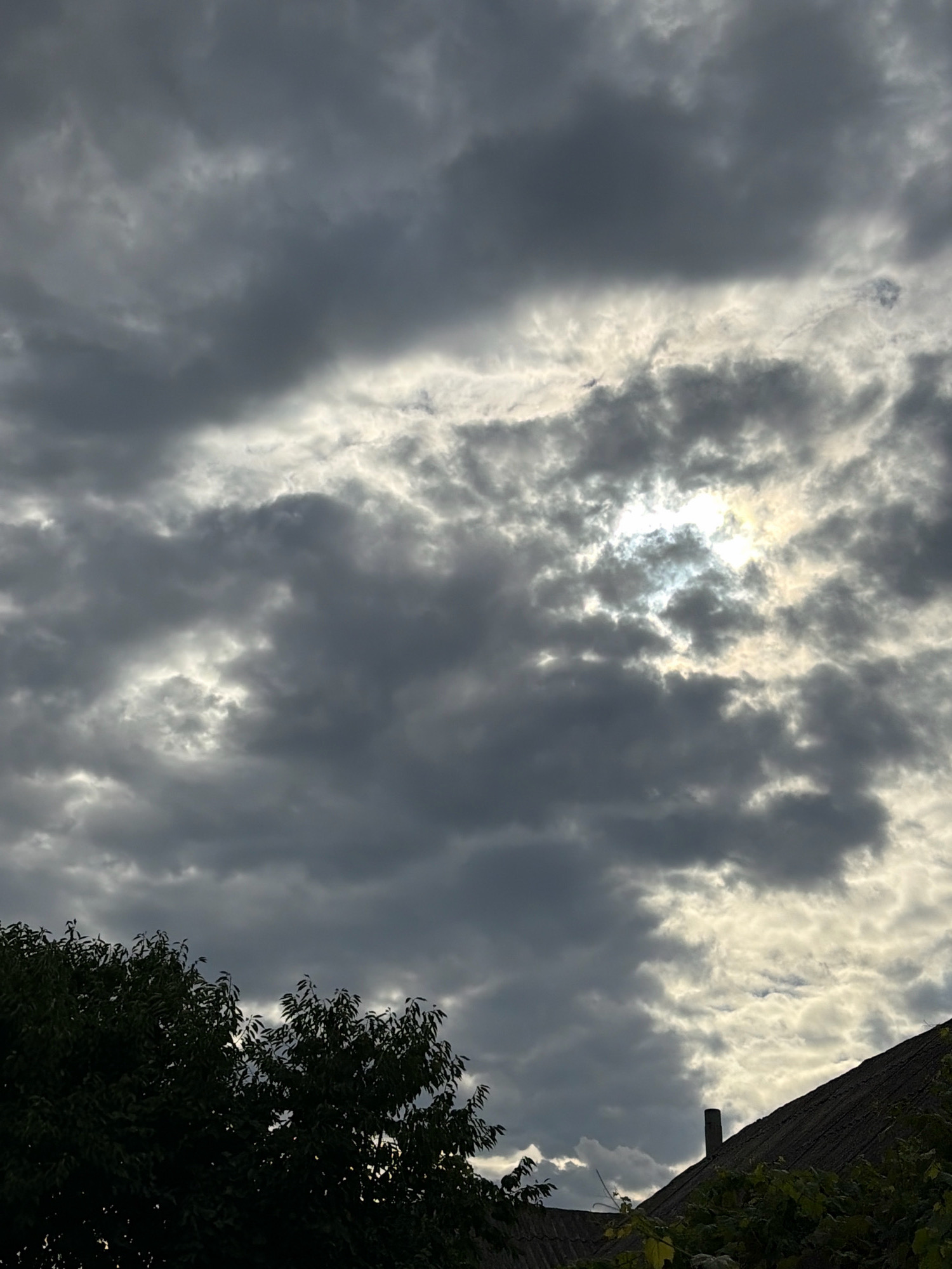 The image displays a sky filled with dark, dramatic clouds, with patches of sunlight breaking through in the upper right portion of the frame. In the foreground, the dark silhouettes of trees and the roof of a building are visible. The overall impression is one of an overcast or stormy sky, with a hint of light offering a dynamic contrast. The location is stated to be Oxentea, Moldova.