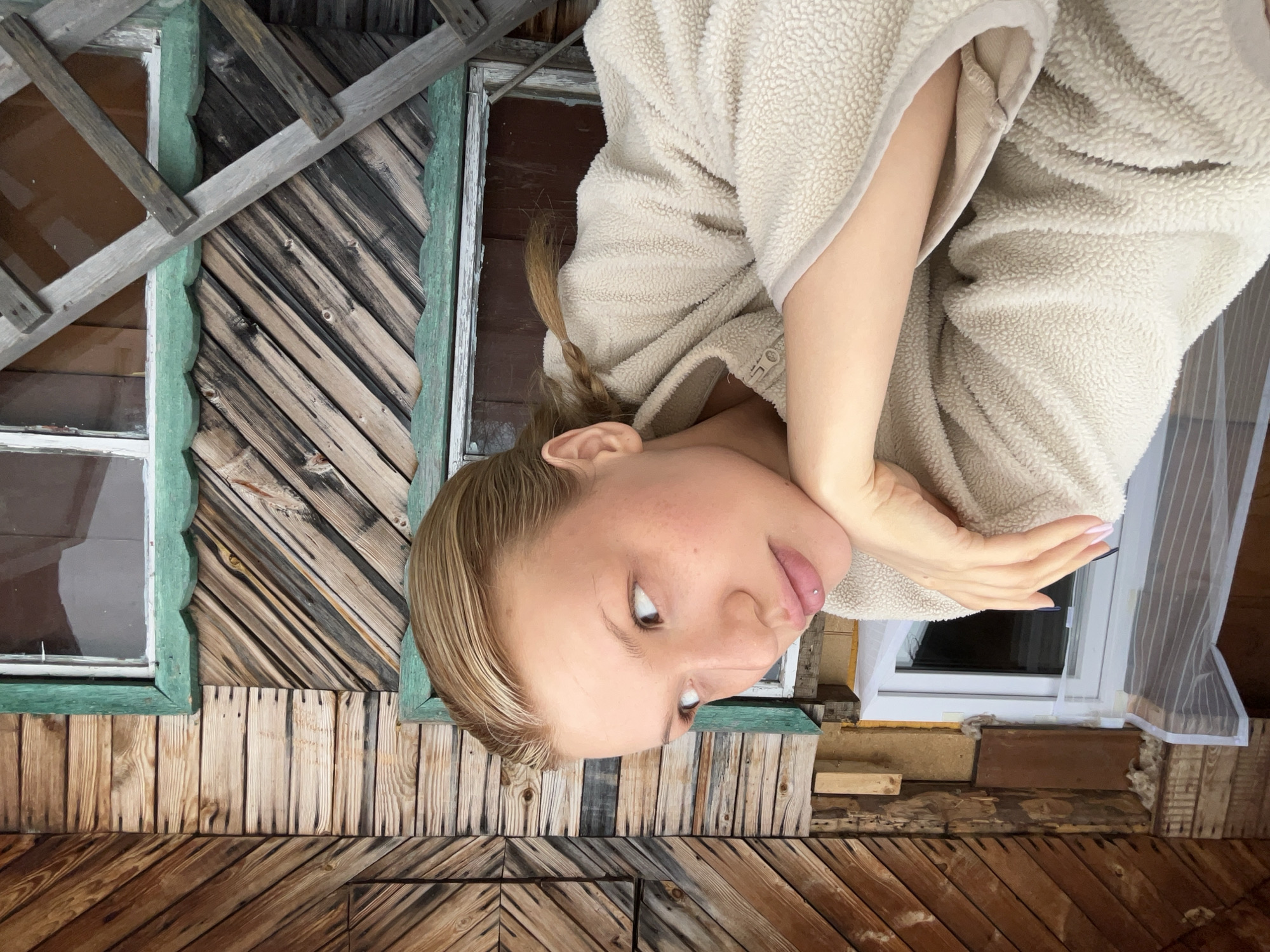A medium shot depicts a woman wearing a beige sherpa jacket. She is situated at an angle, positioned against a weathered wooden structure with two windows. The setting shows an exterior view of what is potentially an old building or house. The windowsills are painted green. This photograph was taken in the city of Chaikovsky, Russia.