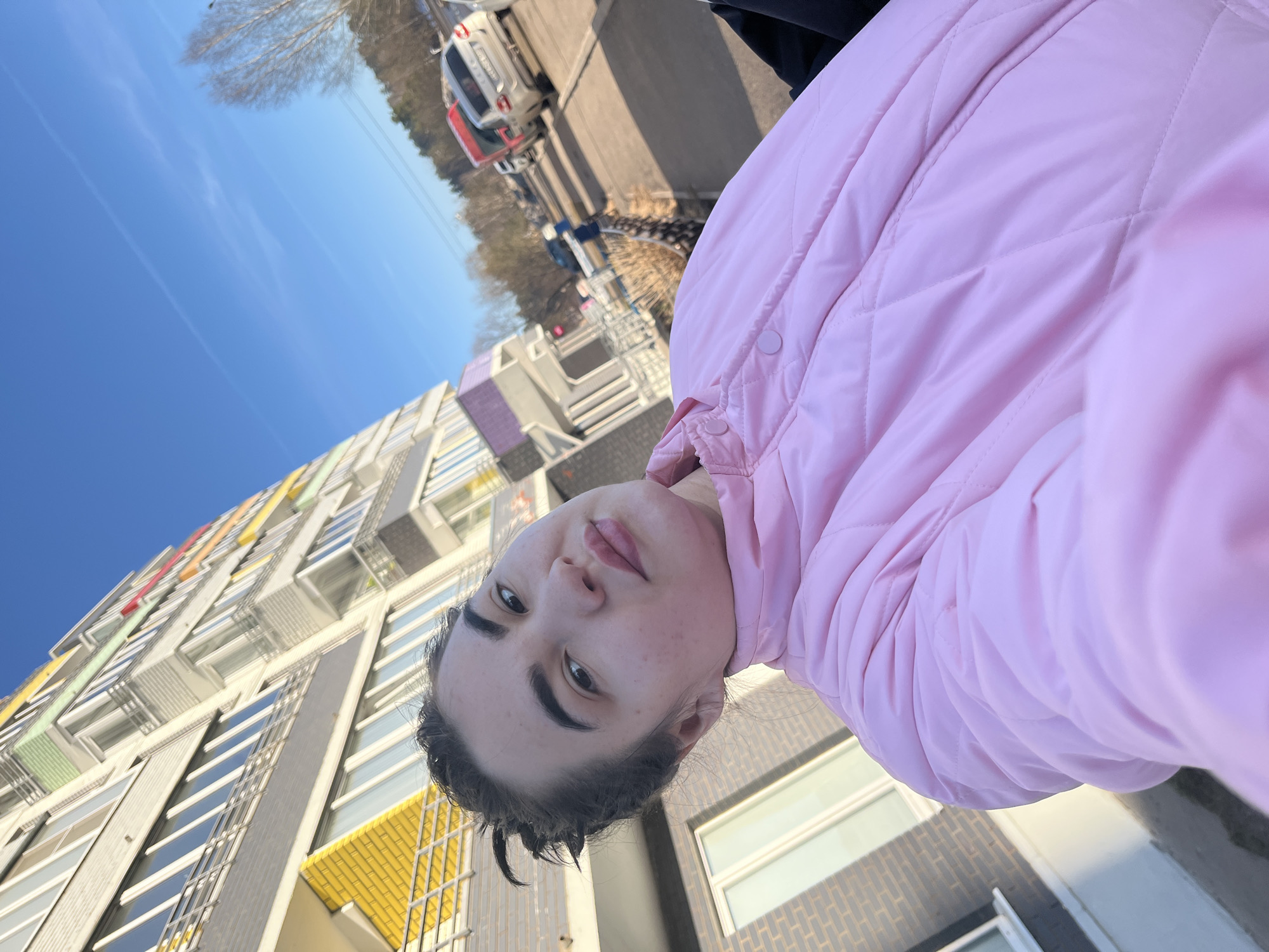 A young woman wearing a pink quilted jacket poses for a selfie outdoors. The background features a multi-story building with a modern design, characterized by colorful rectangular accents on its facade. The clear blue sky is visible behind the building. A road with parked cars and pedestrians is in the distance. Based on the architectural style of the building, the location is Chaikovsky, Russia.