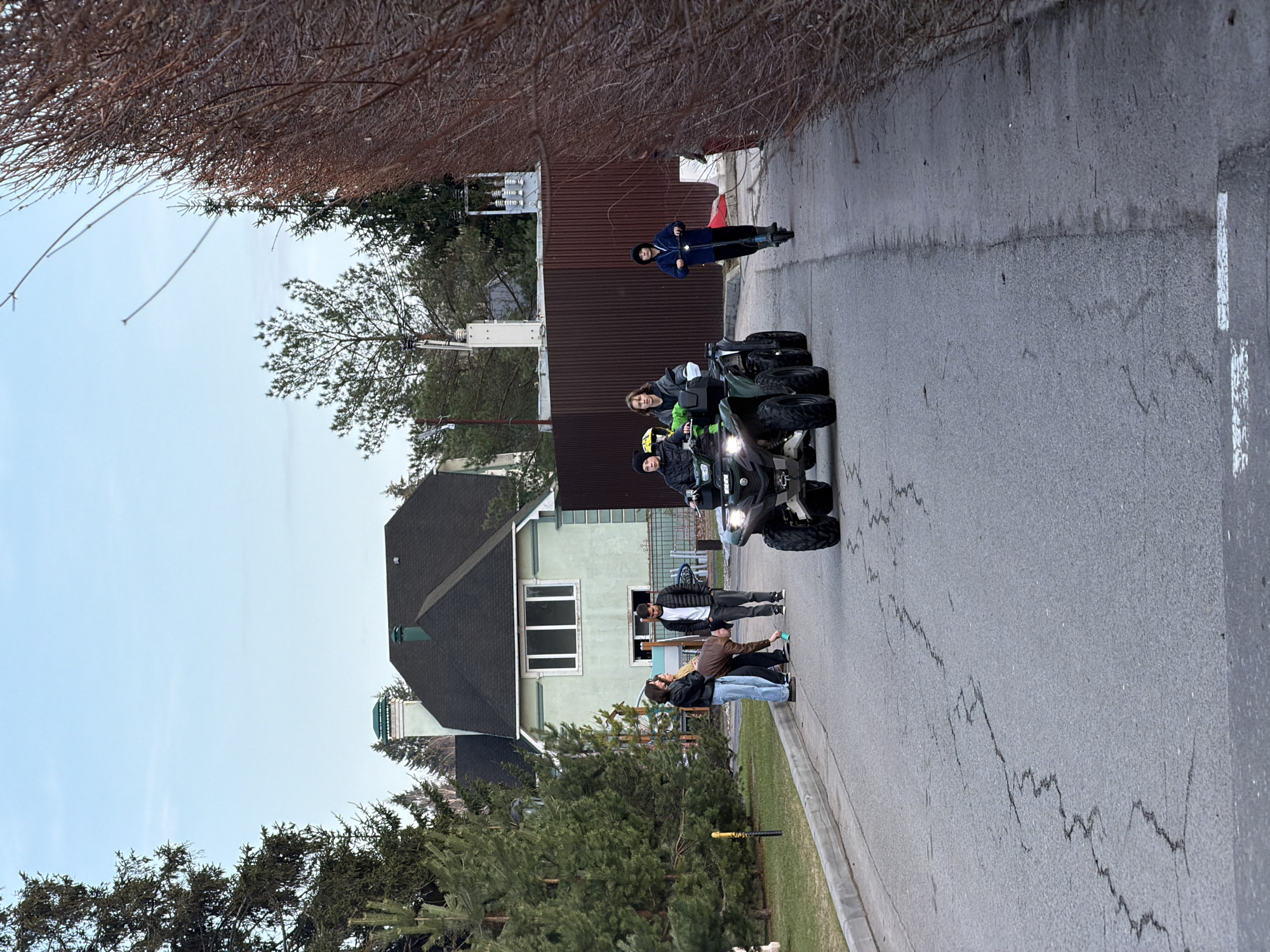 The scene depicts a street in Mishutkino, Russia. Three people are riding two ATVs down a paved road. One ATV appears to be dark green and the other appears to be black. A group of four people are standing along the side of the road observing them. One individual, wearing a blue jacket, is standing farther down the street facing the ATVs. Buildings and trees line both sides of the street. The sky is overcast. There are cracks in the pavement of the road.