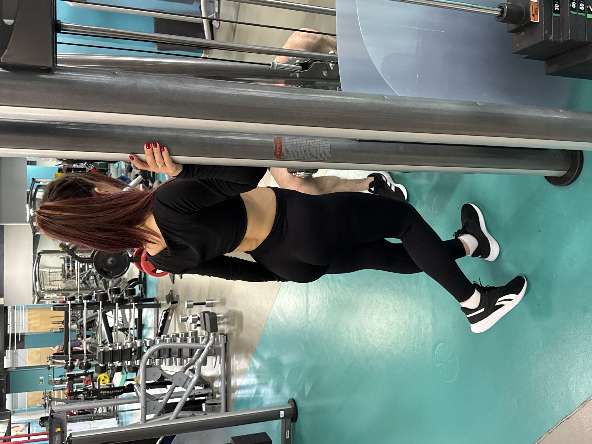 A woman in black workout attire is positioned in a gym, holding onto a piece of gym equipment. She is wearing black leggings, a black long-sleeved crop top, and black-and-white sneakers. Her hair is reddish-brown and pulled back. The gym has a variety of workout equipment, including weight machines and dumbbells. The floor is a light green color. A man can be seen lying on a bench in the background. The location is in the city of Yekaterinburg, Russia.