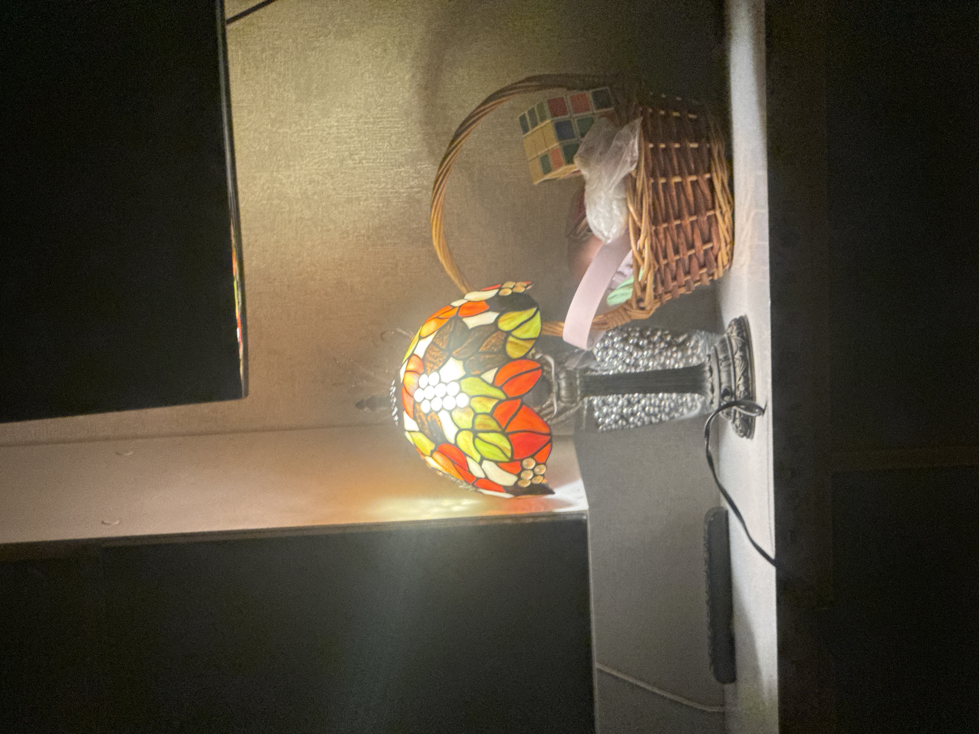 The image shows a dimly lit room with a Tiffany-style lamp on a shelf. Next to the lamp is a woven basket with a Rubik's Cube inside. The location cannot be determined from the image.