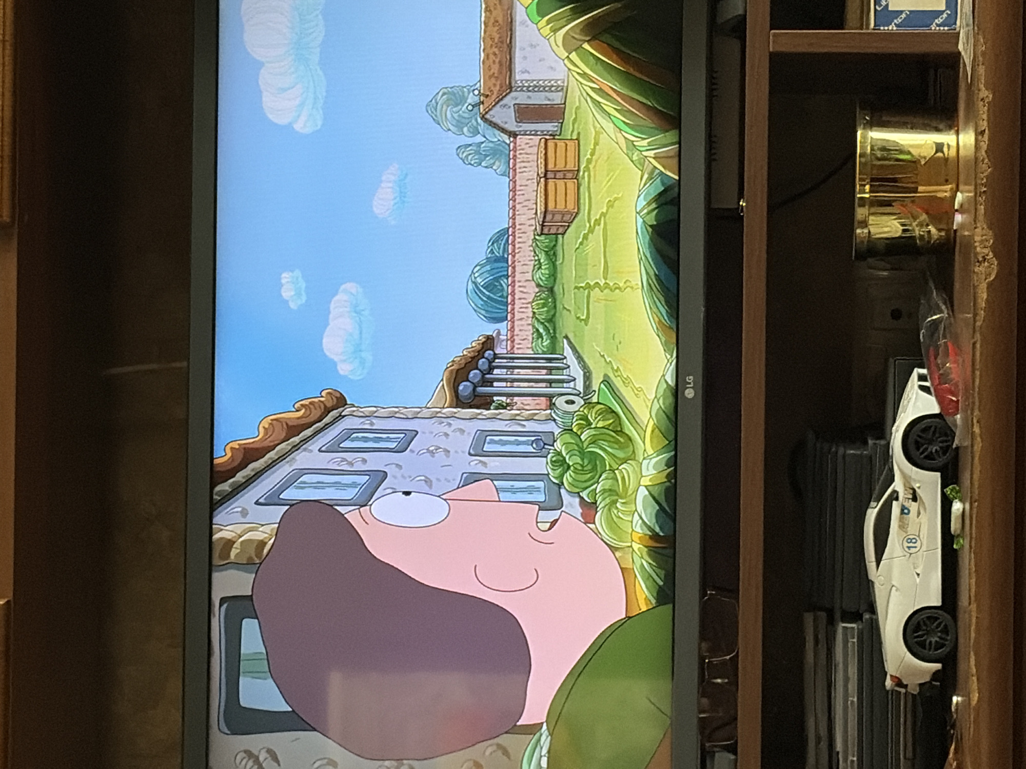 The image displays a television screen showing a scene from an animated show, possibly "Rick and Morty." The animated scene depicts a backyard with a house, a wall, and a garden. On the left side of the screen, a character's face, rendered in a cartoon style with large eyes and brown hair, is prominently featured in the foreground, looking towards the backyard. The backyard itself is green and appears to be well-maintained, with several structures and decorative elements.
Below the television, on a wooden shelf, several objects are visible. These include a white toy sports car, a stack of DVDs or video games, a pair of eyeglasses, a golden cylindrical container, and other assorted items. The overall setting appears to be a room in a residential home.