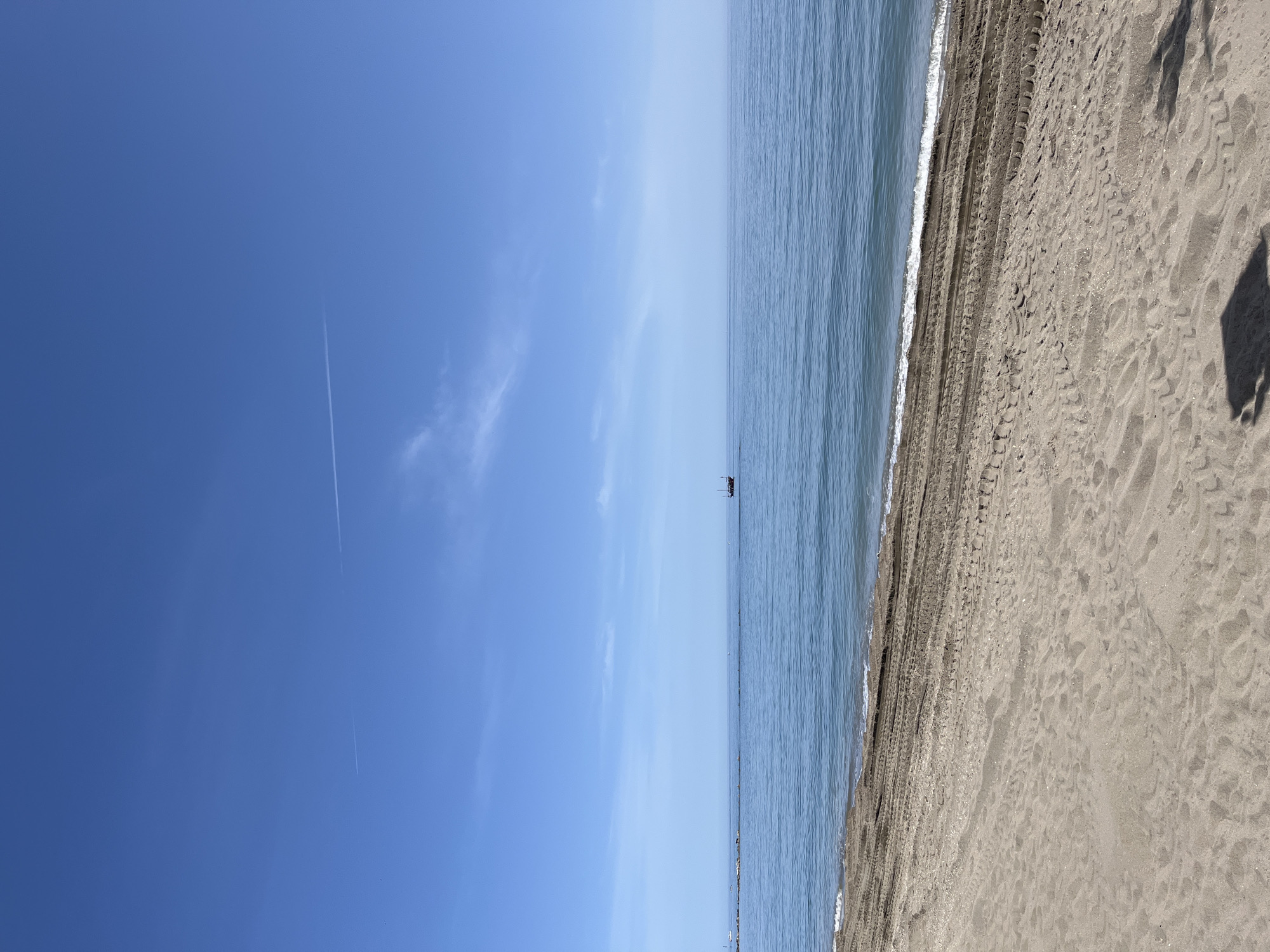 The image captures a serene coastal landscape on a clear, sunny day. The scene is horizontally divided into three main sections: sky, sea, and beach.
The upper portion of the image is dominated by a vast, vibrant blue sky. A faint, white contrail from an aircraft is visible, stretching diagonally across the sky. There are also a few wispy, light white clouds scattered sparsely.
The middle section features the calm, light blue sea, extending to the horizon. Gentle ripples and small waves are noticeable closer to the shore. Far out on the water, near the horizon, a small, dark, rectangular object, possibly a boat or a buoy, can be seen.
The bottom section of the image shows a wide expanse of light brown/tan sand. Distinct tire tracks or other vehicle marks run parallel to the waterline. Closer to the water, the sand appears darker due to moisture from the gentle waves. Numerous footprints are also visible on the dry sand. In the bottom right corner, a large, dark, irregularly shaped shadow is cast on the sand, likely belonging to the photographer or an object nearby.
The bright sunlight suggests it is either morning or early afternoon. The overall atmosphere is one of tranquility and natural beauty. No discernible text is visible within the image. The location context indicates this scene is at Arroyo de la Miel-Benalmádena Costa, Spain.