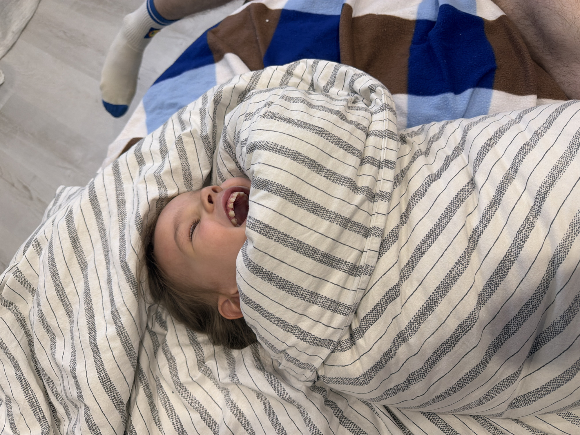 A young child lies wrapped in a striped blanket, their head emerging at the top, mouth open, possibly laughing or shouting. Part of a checkered blanket with brown, blue, and light blue squares is visible behind the child. A person wearing a sock with a blue toe and blue stripes is at the top left. Another person's leg is visible at the top right. The setting seems to be indoors. There is no evidence to connect the image with the city of Chaikovsky, Russia.