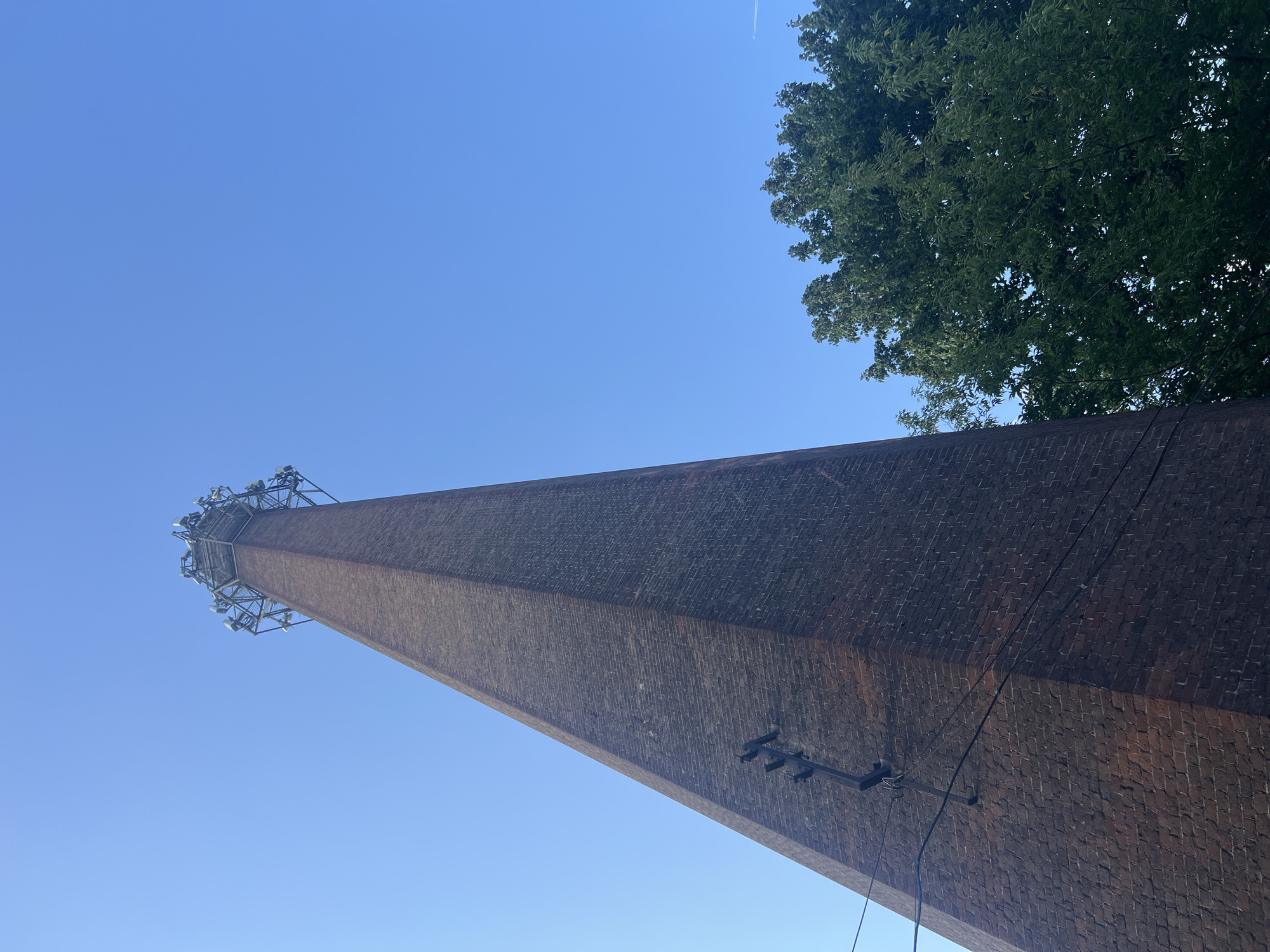 The image captures a low-angle view of a tall, brick chimney against a clear blue sky. The chimney tapers upwards and is topped with a metallic structure holding numerous antennas, suggesting it functions as a telecommunications tower. There are power lines running along the side of the chimney, supported by a wooden pole attached to the brickwork. To the right of the chimney, a dense green tree is visible, its foliage reaching towards the sky. The scene appears to be outdoors, likely in an urban or semi-urban environment. There are no people or animals depicted in the image.