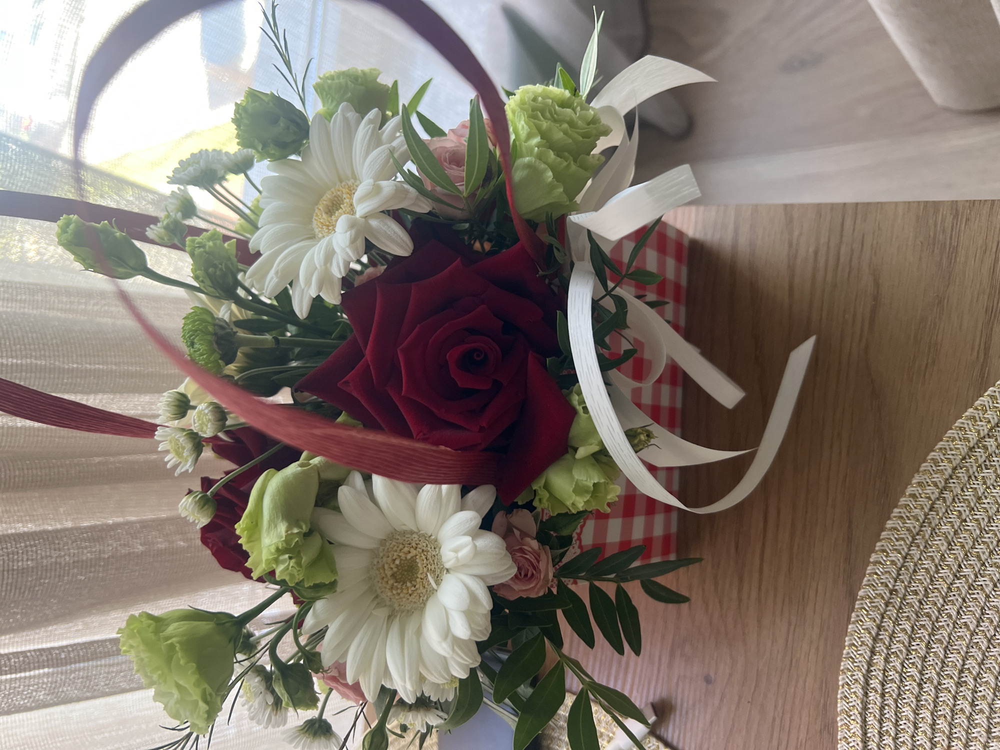 The image shows a close-up of a floral arrangement presented in a red and white checkered box. The arrangement features a prominent deep red rose at its center, surrounded by white gerbera daisies, light green eustomas, and small green carnations. There are also delicate white aster-like flowers and green foliage. White ribbons are tied around the box, and a red ribbon accent is visible within the arrangement. The box sits on a wooden surface, with a woven placemat visible in the lower left corner. In the background, a sheer curtain hangs, and light streams in, creating a bright, slightly hazy atmosphere. The context provided indicates the scene is located in the city of Plovdiv, Bulgaria, though the specific location within the city is not visually evident.