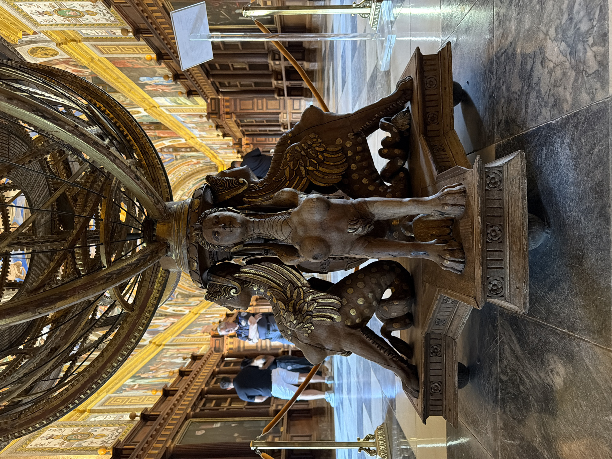 The image shows the armillary sphere in the Royal Library of San Lorenzo de El Escorial, Spain. The sphere is supported by a wooden pedestal adorned with carved mythological figures, including a sphinx-like creature in the center. The library's interior is visible in the background, featuring ornate wooden shelves lining the walls, decorated with a frieze of paintings. Two individuals are standing near the sphere, looking at it. The floor is a checkered pattern of marble tiles.