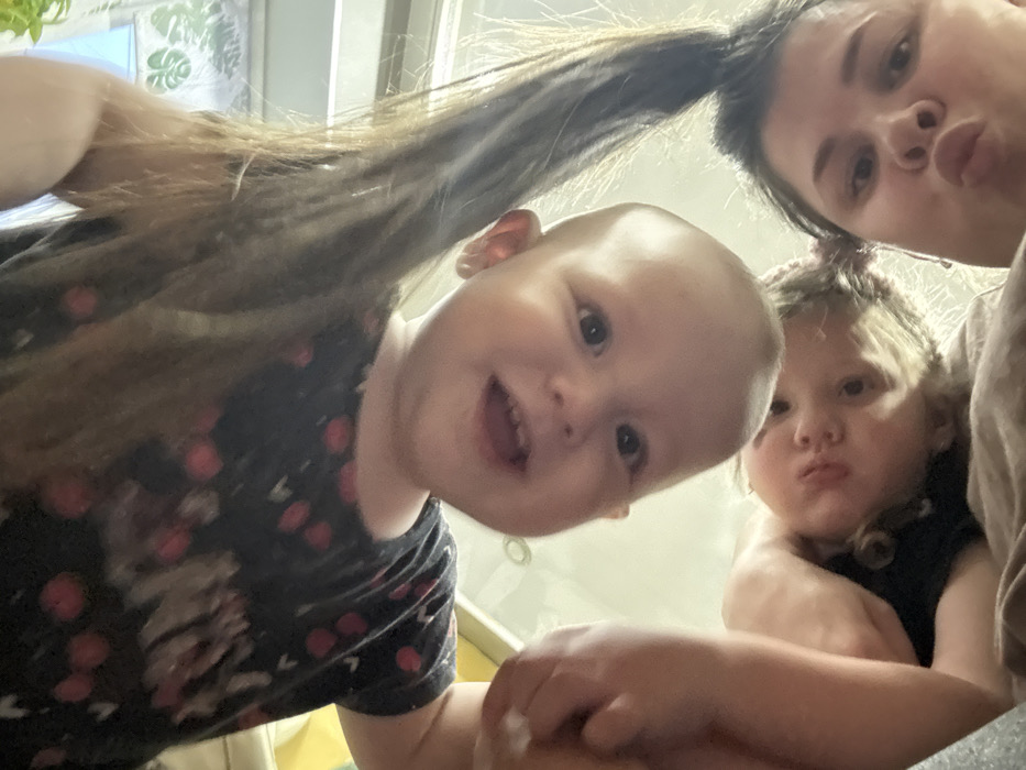 This image, captured in the city of Кипчак, Russia, presents a low-angle, inverted view of three individuals looking downwards into the camera. At the top of the frame, centered, is an infant or young toddler with light skin, dark eyes, and an open mouth revealing upper teeth, wearing a dark top with red and white circular patterns. Long, dark hair cascades downwards on either side of the infant's head, likely belonging to an adult situated behind. To the left and slightly below the infant is a second, slightly older child with light, curly hair and pouting lips, wearing a dark top. In the lower right quadrant, a female adult with dark hair and pouting lips looks directly into the camera. The background is softly blurred and brightly lit, with a faint green plant motif visible in the upper right.