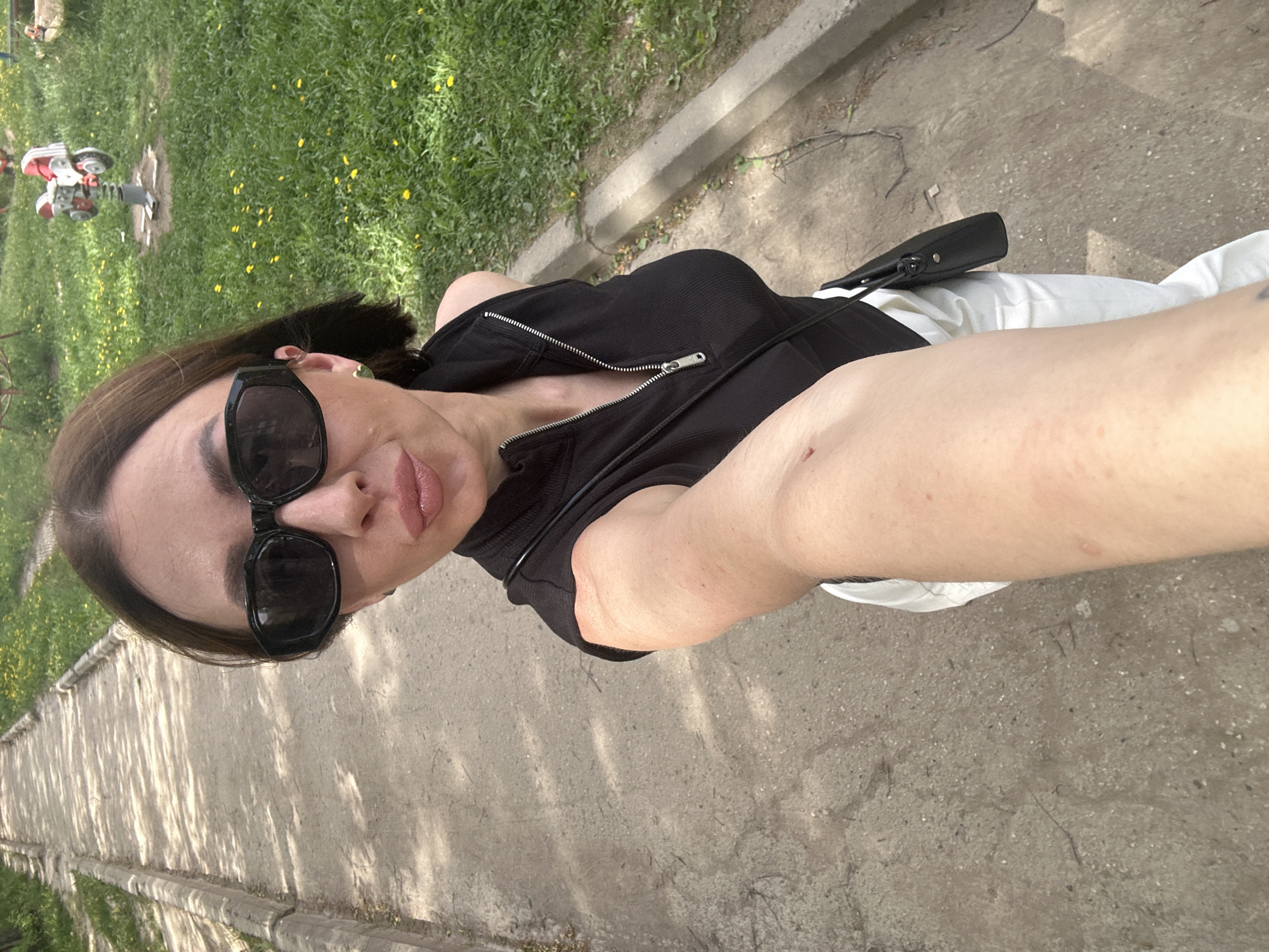 A selfie taken at ground level depicts a woman in sunglasses standing on a concrete path, likely in Votkinsk, Russia. The woman is wearing a black zipped top and white trousers. She has dark hair and light skin. To the left of the path is a grassy area with dandelions and a child's spring rider toy. The path is bordered by a low concrete curb.